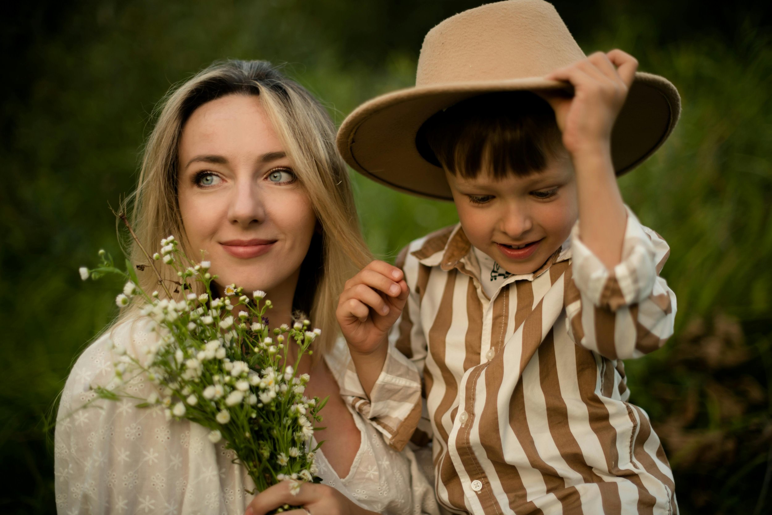 A woman with blonde hair and blue eyes holding a bouquet of white daisies, standing beside a young boy with dark hair wearing a beige hat and a striped shirt, both outdoors with green foliage in the background.