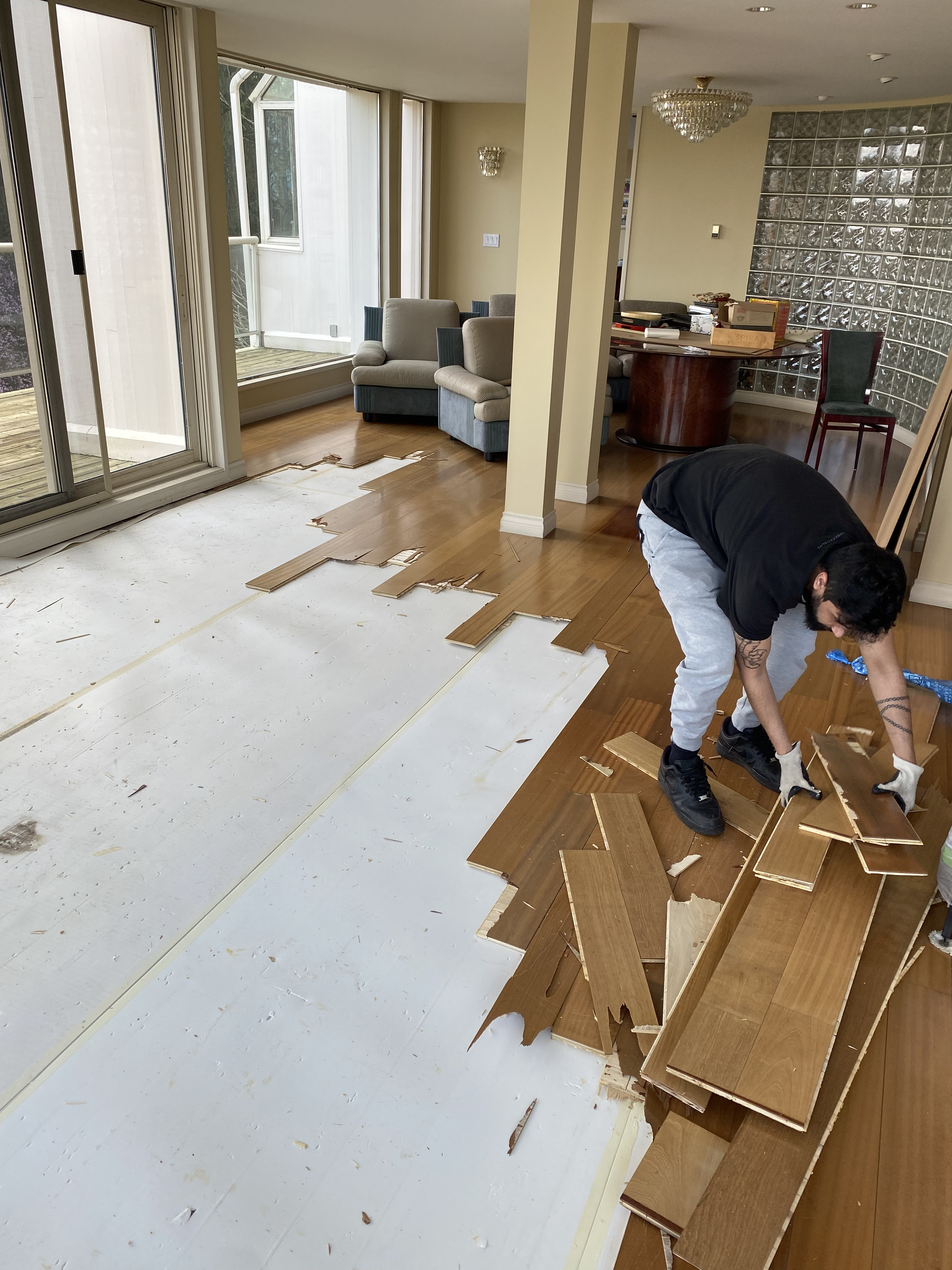 A person is removing a damaged wooden floor in a living room with large windows, beige walls, a chandelier, and modern furniture.