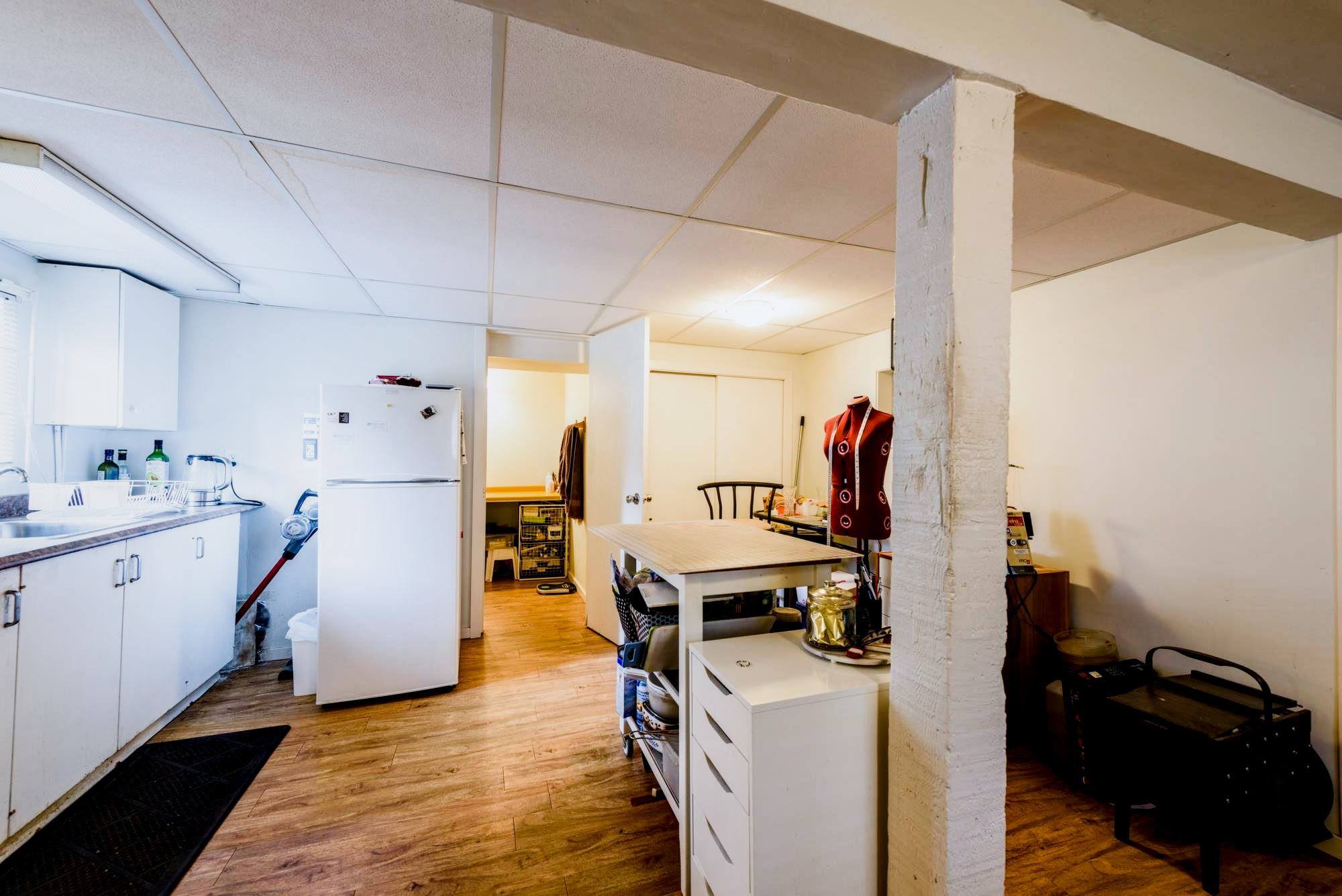 A small kitchen and dining area with wood floors, white cabinets and appliances, and a white table with a small dresser or storage unit nearby. There is a decorative mannequin torso, a black chair, and various small items on the furniture. An open doorway leads to another room, and the ceiling has square tiles.