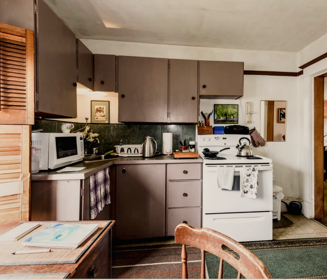 A small kitchen with dark cabinets, a white stove with a kettle, a microwave, and various kitchen items on the countertop, a wooden chair, and a small table with books.