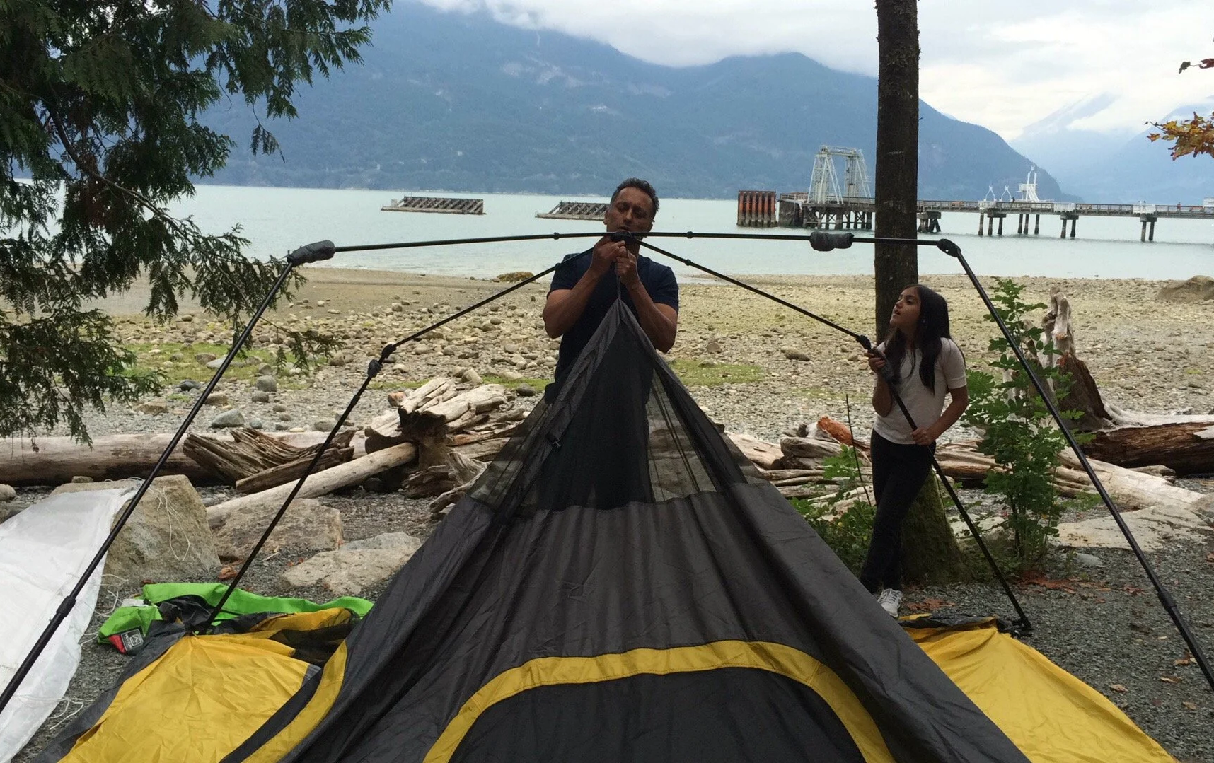 A man and woman setting up a camping tent on a rocky beach with a lake and mountains in the background, near a pier extending into the water.
