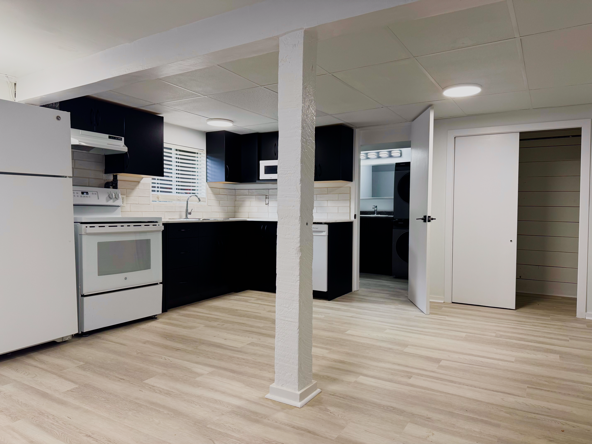 A kitchen with white appliances and black cabinets, a white brick backsplash, a window with blinds, and a metal microwave. An open door reveals a laundry area with a washer and dryer.