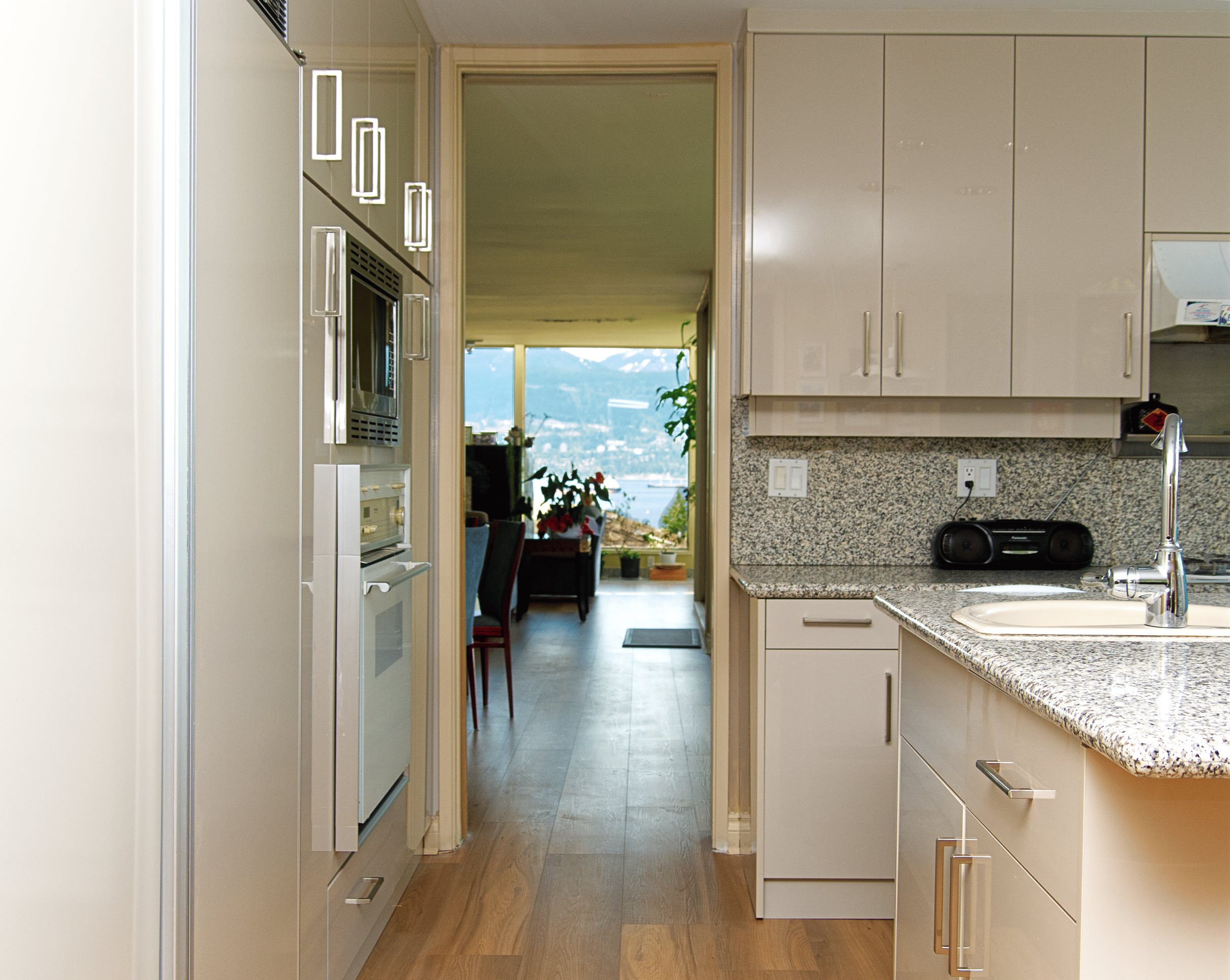 View of a modern kitchen with white cabinets and granite countertops, a view into a dining area with a window overlooking a body of water and mountains, and hardwood flooring throughout.