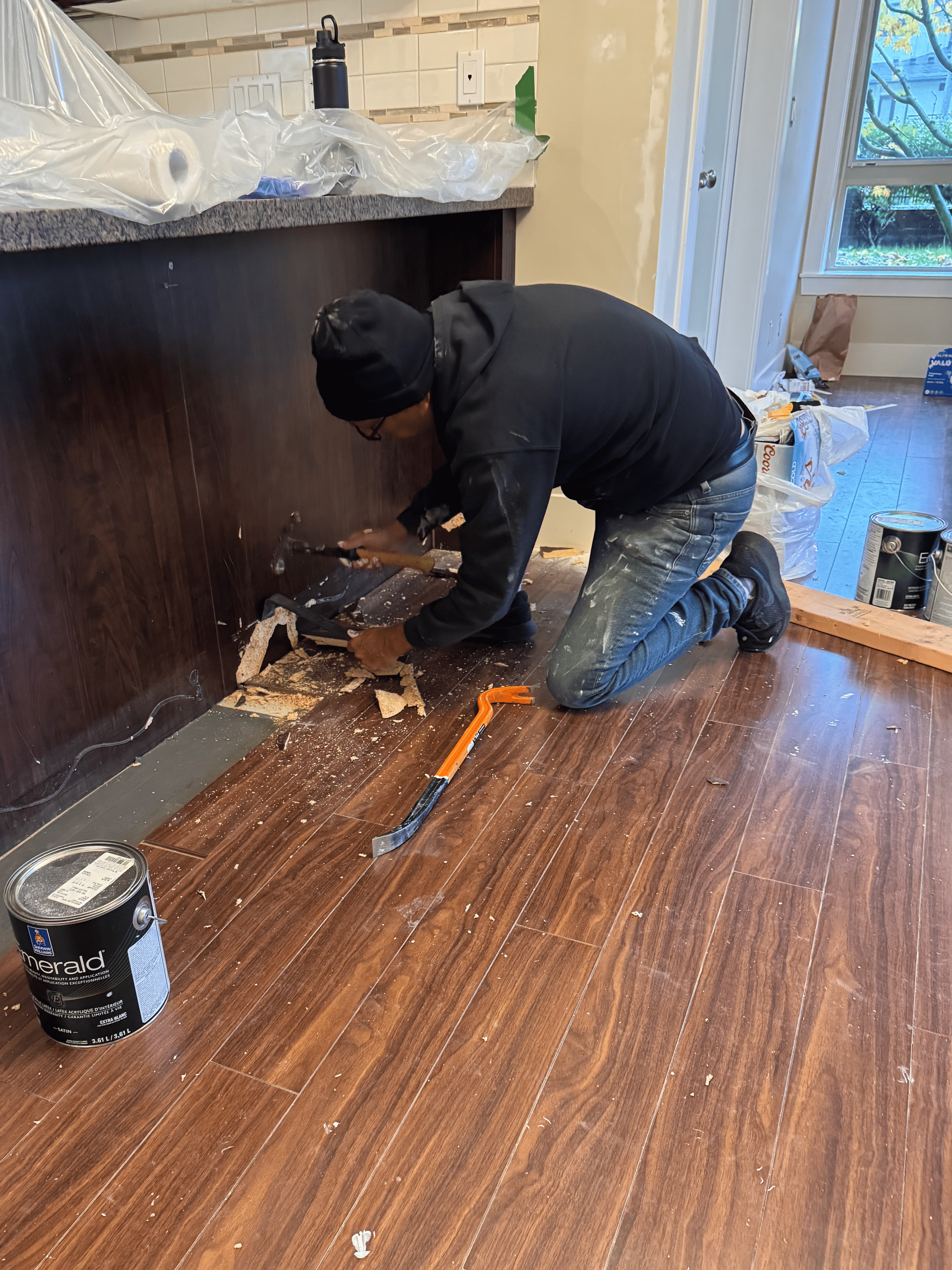 A person kneeling on the floor, demolishing a kitchen cabinet, with tools and debris around, during home renovation.