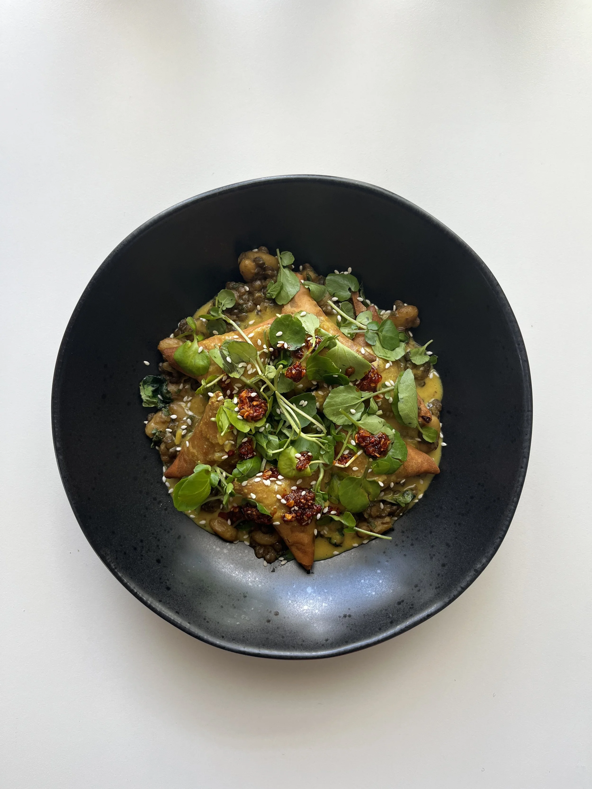 A black bowl of Asian dumplings topped with herbs, sun-dried tomatoes, sesame seeds, and microgreens, on a white surface.