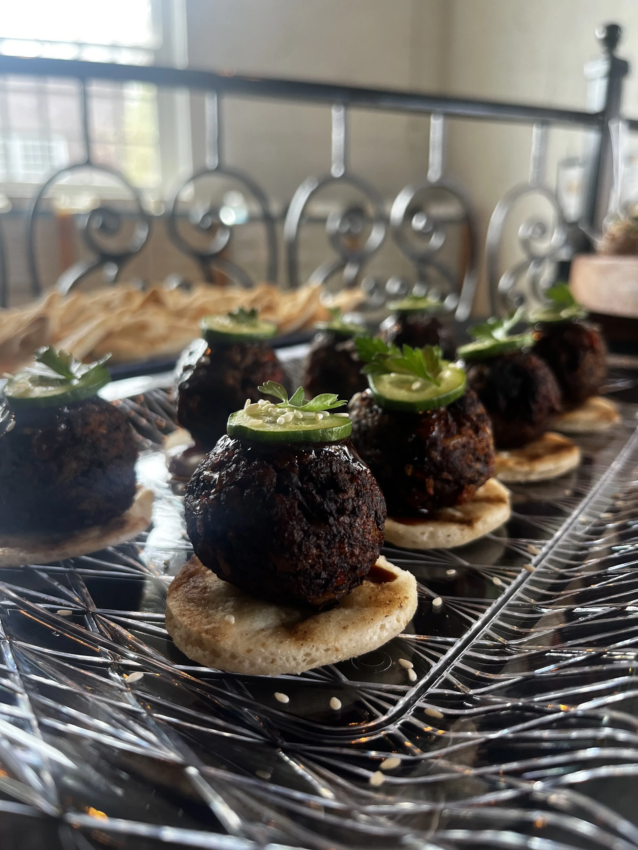 Close-up of small appetizers on a wire cooling rack, consisting of round crackers topped with dark meatballs, cucumber slices, and garnished with parsley and sesame seeds.