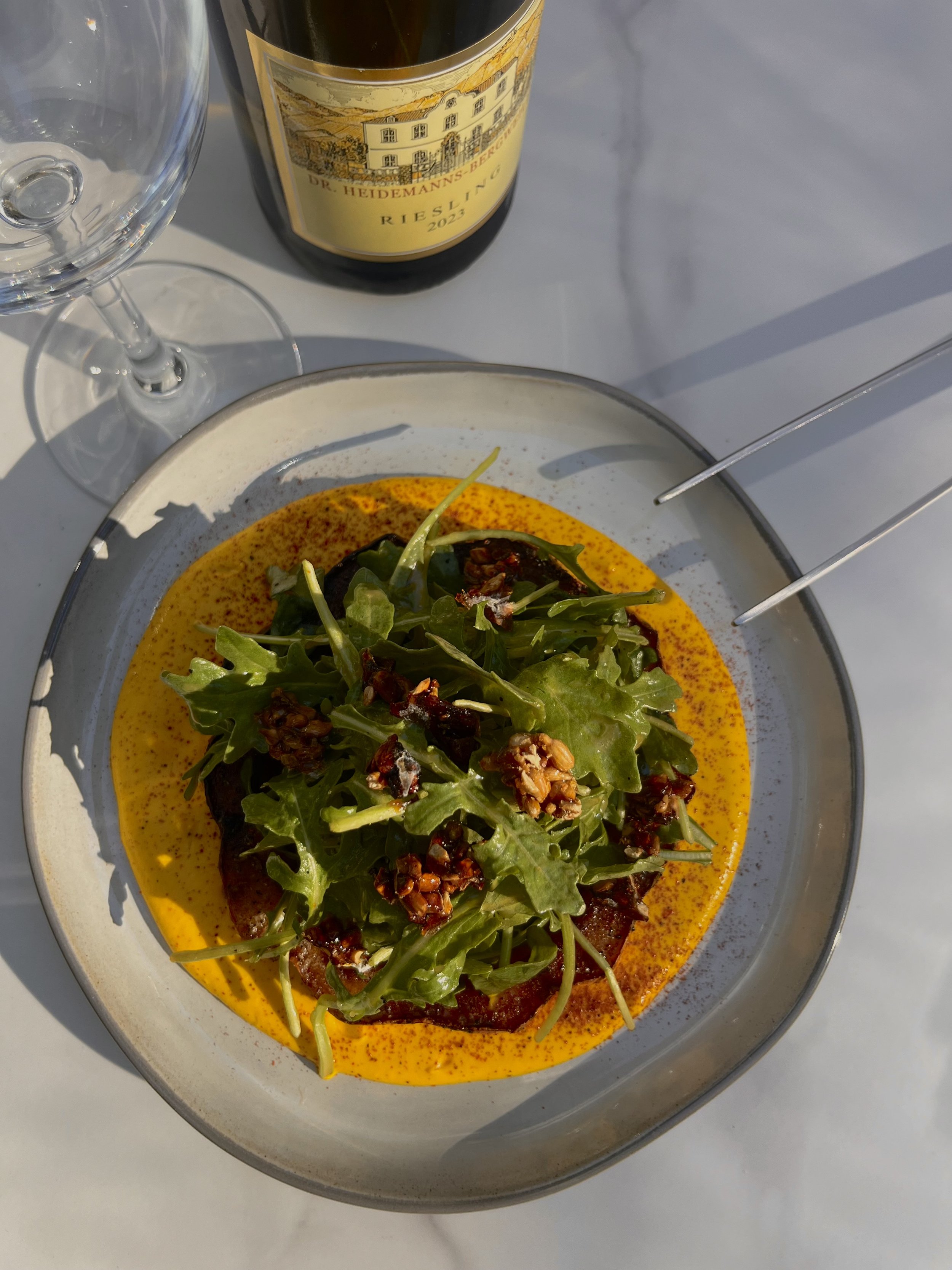 A plate with a salad topped with greens and nuts, surrounded by a yellow sauce or dressing. In the background, there is a bottle of Riesling wine and a partially visible empty wine glass on a white surface.