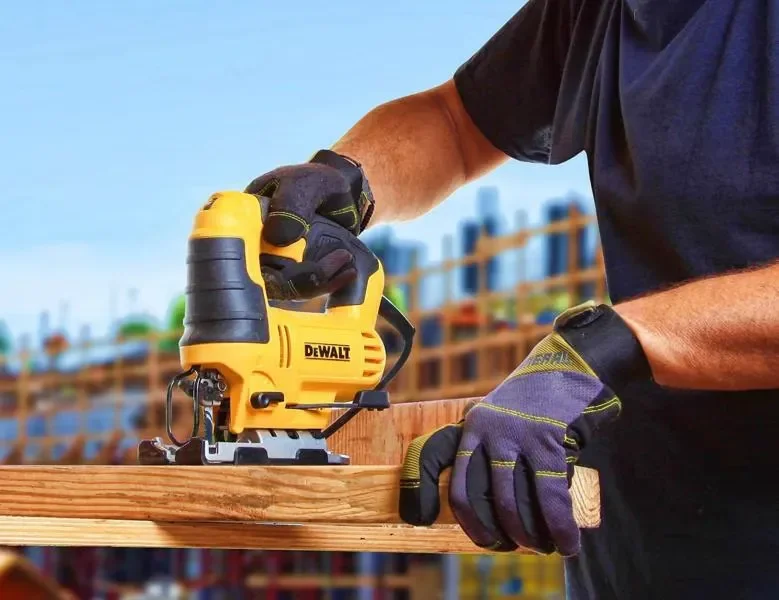 Persona usando guantes de trabajo, manejando una sierra de calar amarilla marca DeWalt para cortar madera en un sitio de construcción.
