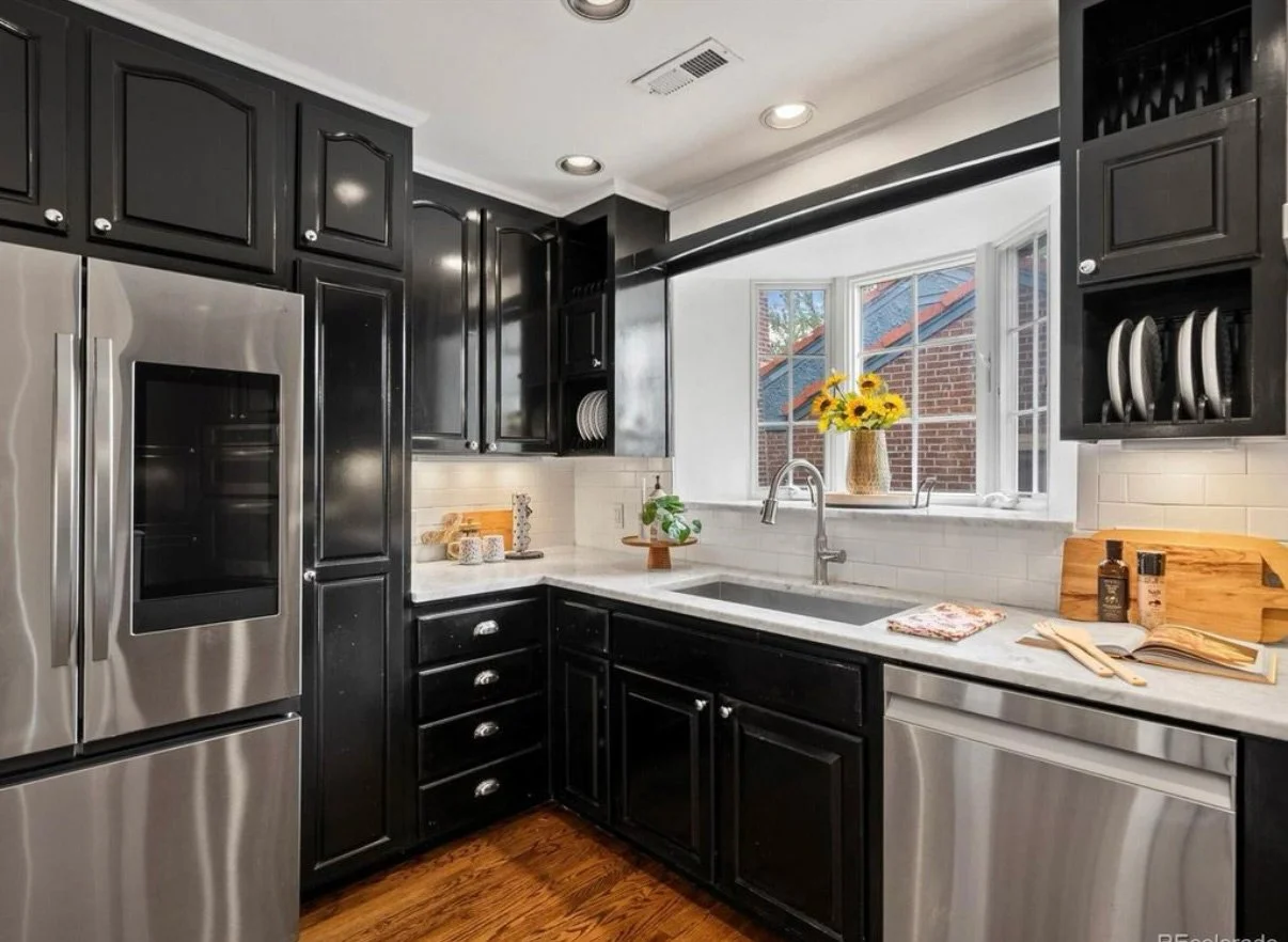 Modern kitchen with black cabinets, stainless steel appliances, a white subway tile backsplash, a window with a view of a brick building, and decorative items including sunflowers, a plant, and kitchenware.