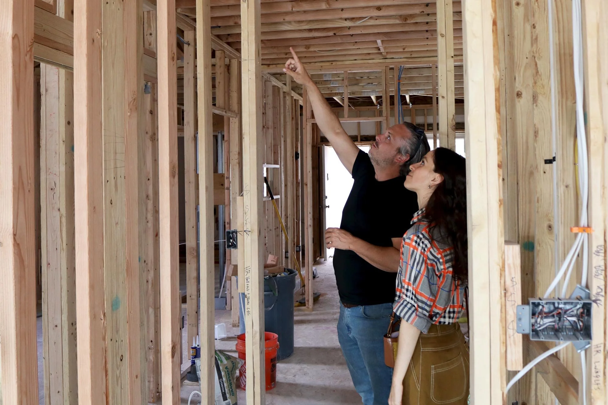 A man and woman look up and point at the unfinished ceiling of a house under construction, surrounded by exposed wooden framing and electrical wiring.