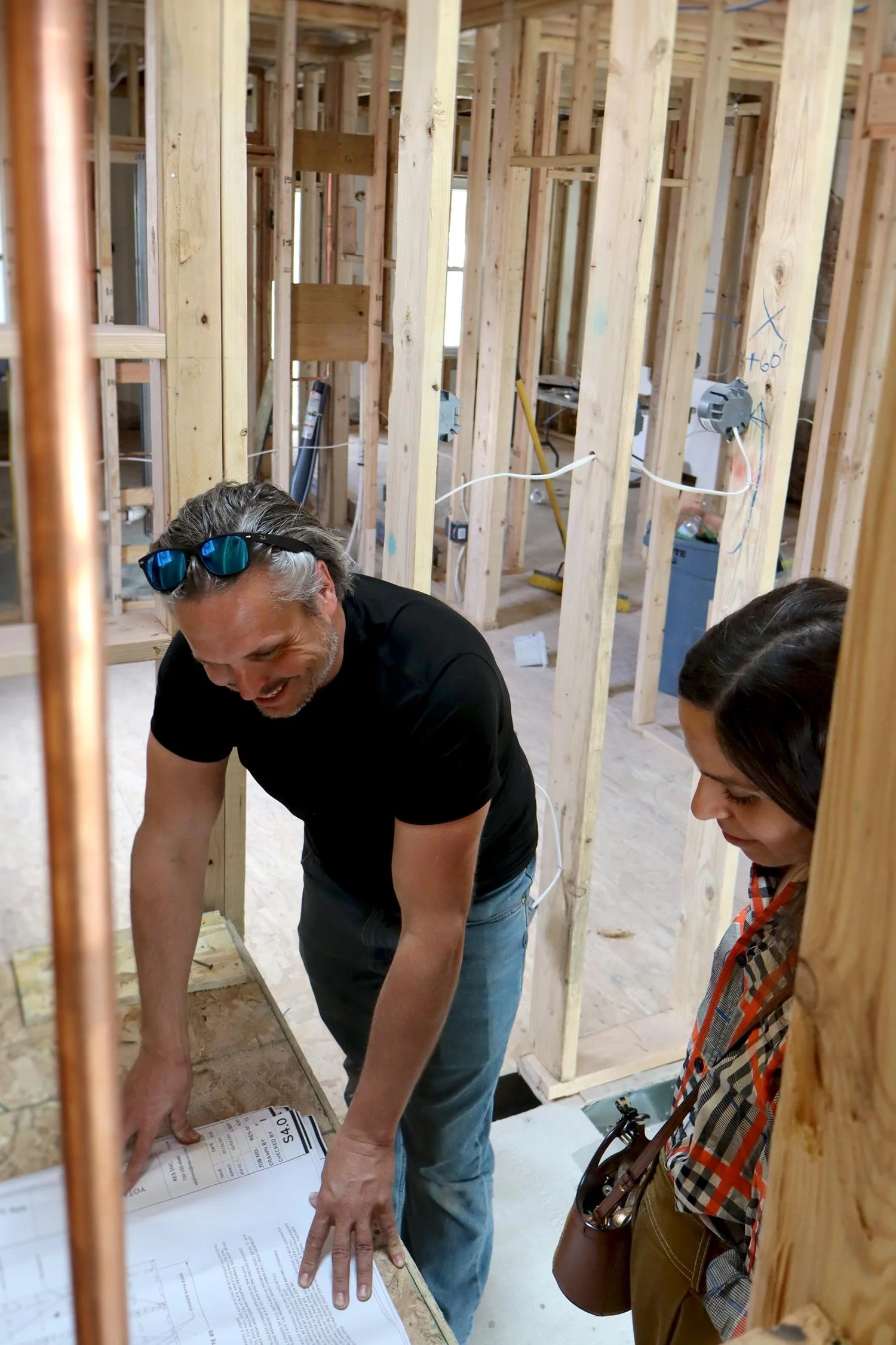 Two people discussing blueprints inside a building under construction with exposed wooden framing, electrical wiring, and construction tools.