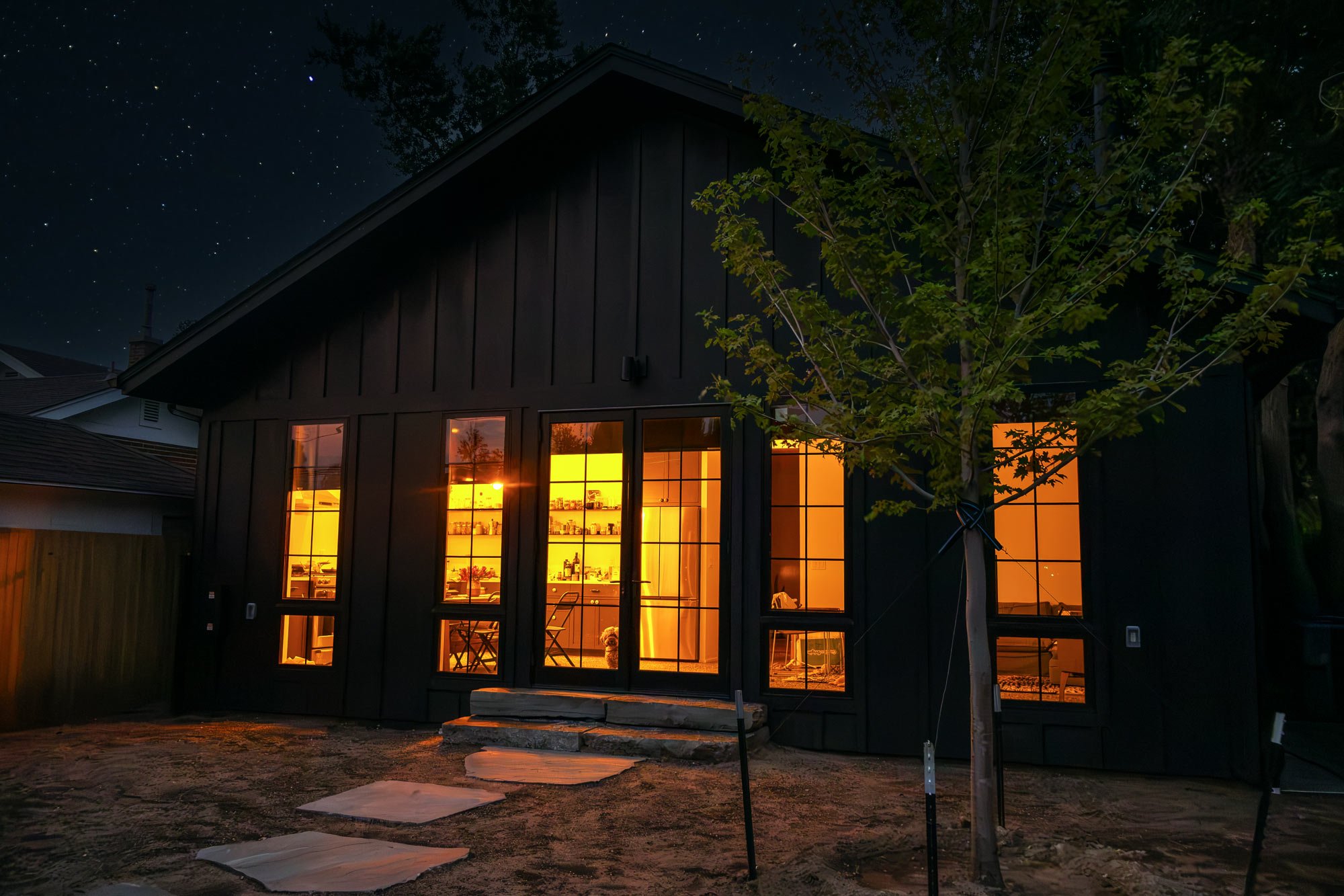 Night view of a house under construction with warm illuminated interior, large windows, a young tree in the foreground, and a starry sky above.