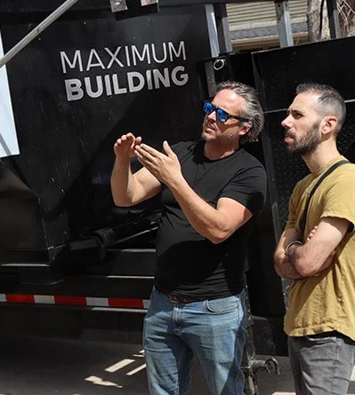 Two men standing outdoors near a black truck with a sign reading 'Maximum Building'. One man is gesturing with his hands, and the other is listening with crossed arms.