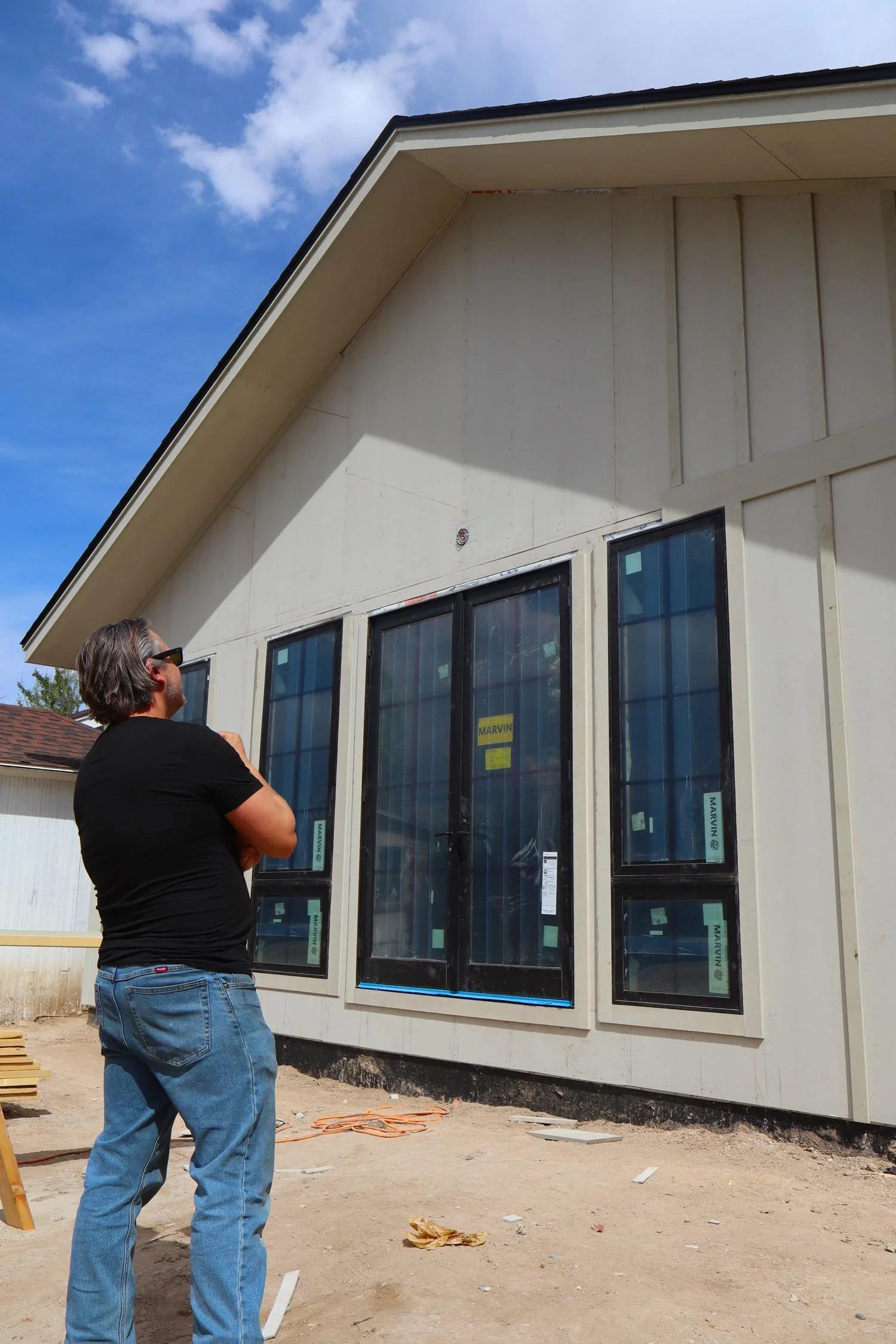 A man in a black t-shirt and blue jeans stands outdoors on a construction site, looking at a house with large window frames. The house has a white exterior, and the roof overhangs with visible construction materials around. The sky is partly cloudy.