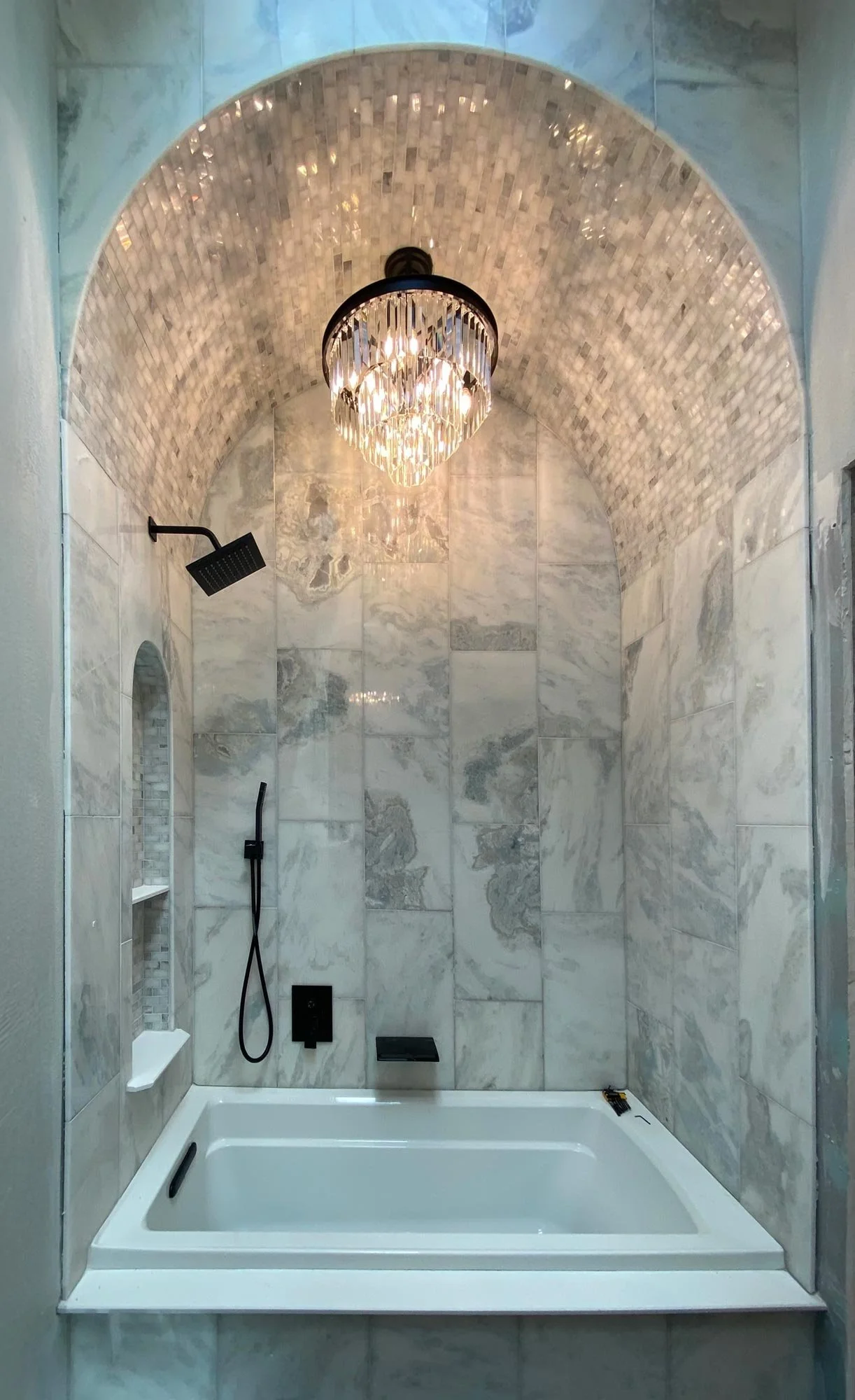 Modern shower with a marble tiled wall, black fixtures, a chandelier hanging from a vaulted ceiling, and a white bathtub at the bottom.