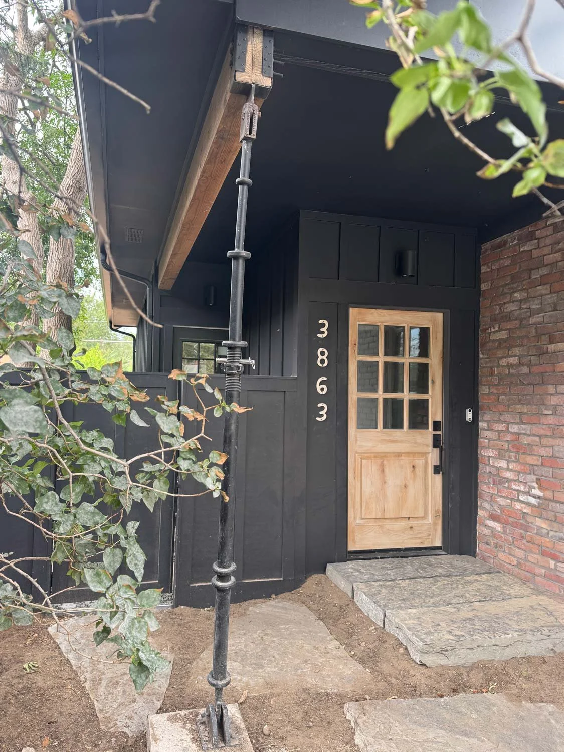 Exterior view of a house with black siding, a wooden door with glass panels, and the house number 3863 displayed vertically beside the door. The porch has a stone step and soil in front. There is a tree branch with leaves partially visible on the left side of the image.