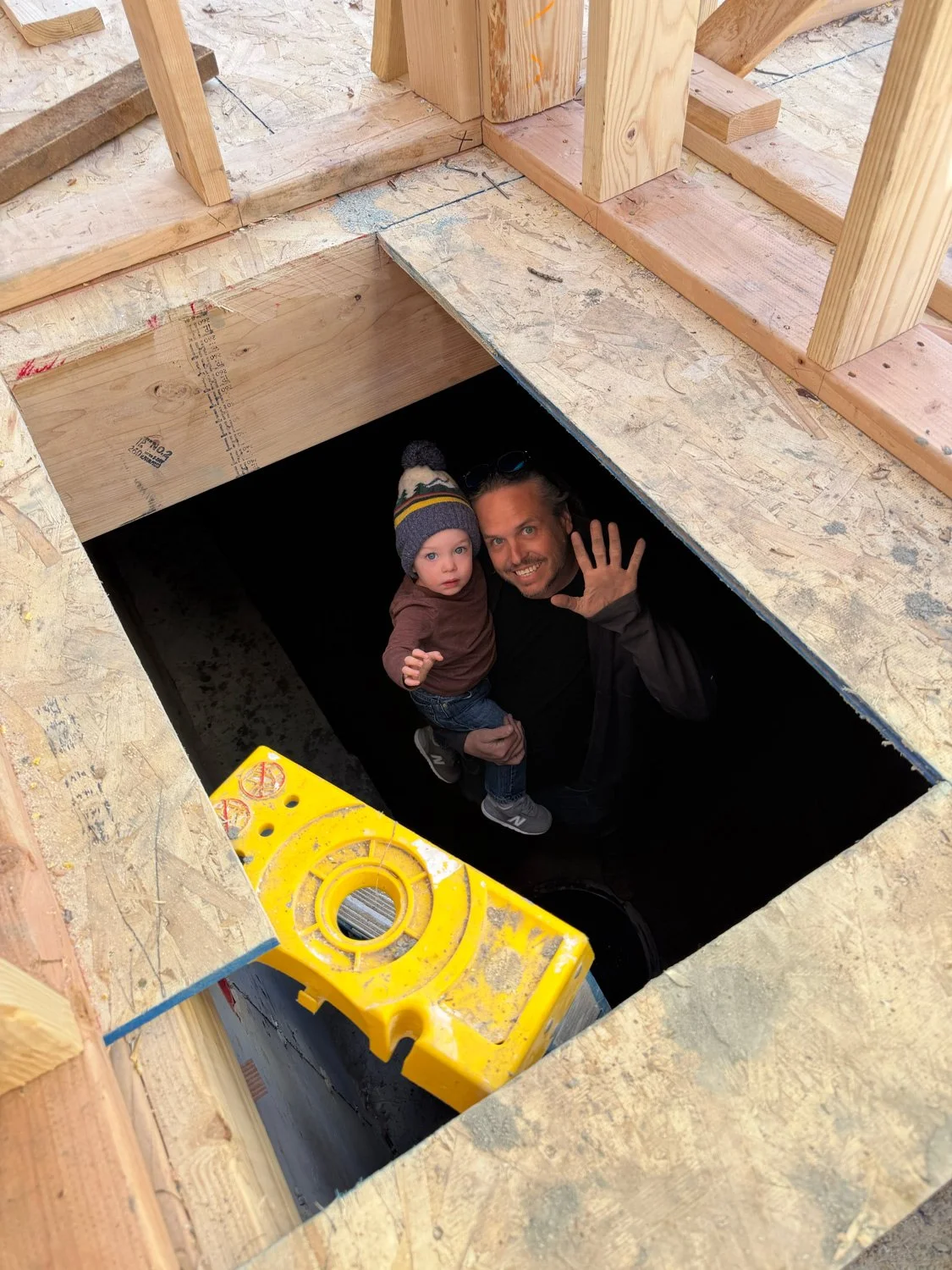 A man holding a young child inside an open construction hole in a building frame, waving at the camera.