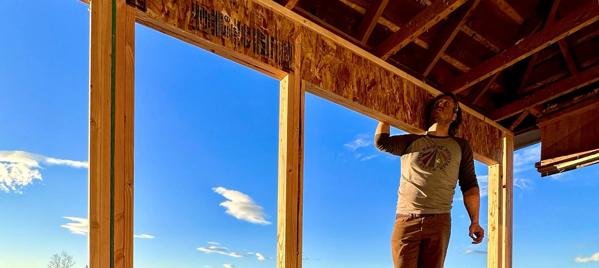 A construction worker in a beige shirt and brown pants inspects the ceiling of a framed building during the daytime with a clear blue sky.