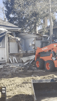 Construction site with a small orange skid-steer loader and a partially demolished house, dirt ground and trees in background.