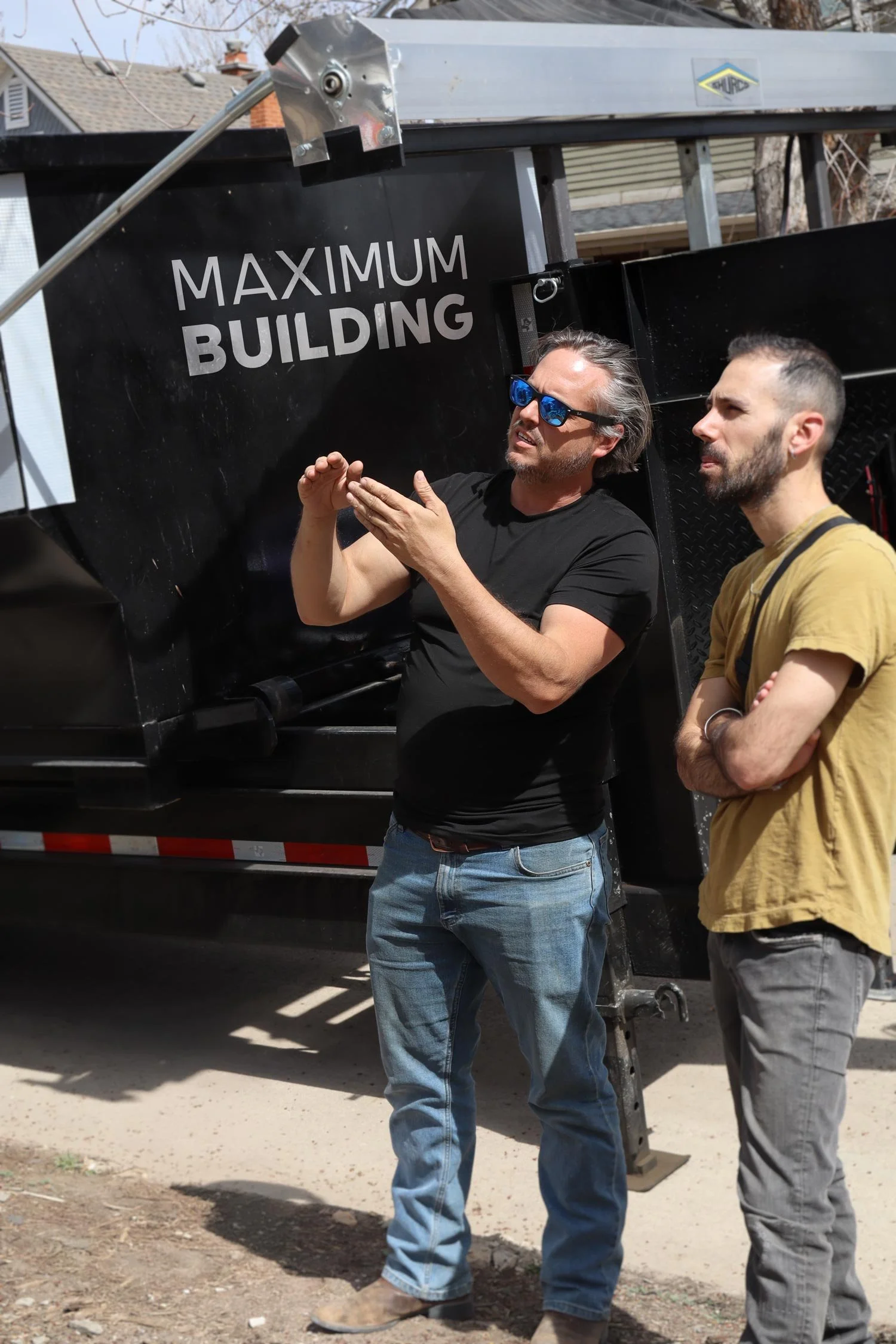 Two men standing beside a flatbed trailer with a sign that reads "Maximum Building". One man wears sunglasses, a black t-shirt, and jeans, while the other wears a yellow t-shirt and gray jeans. They are outdoors in a residential area.