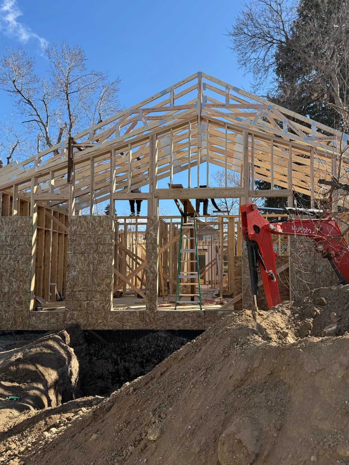 Wooden framing of a house under construction with visible roof trusses, surrounded by dirt and construction equipment, under a clear blue sky.