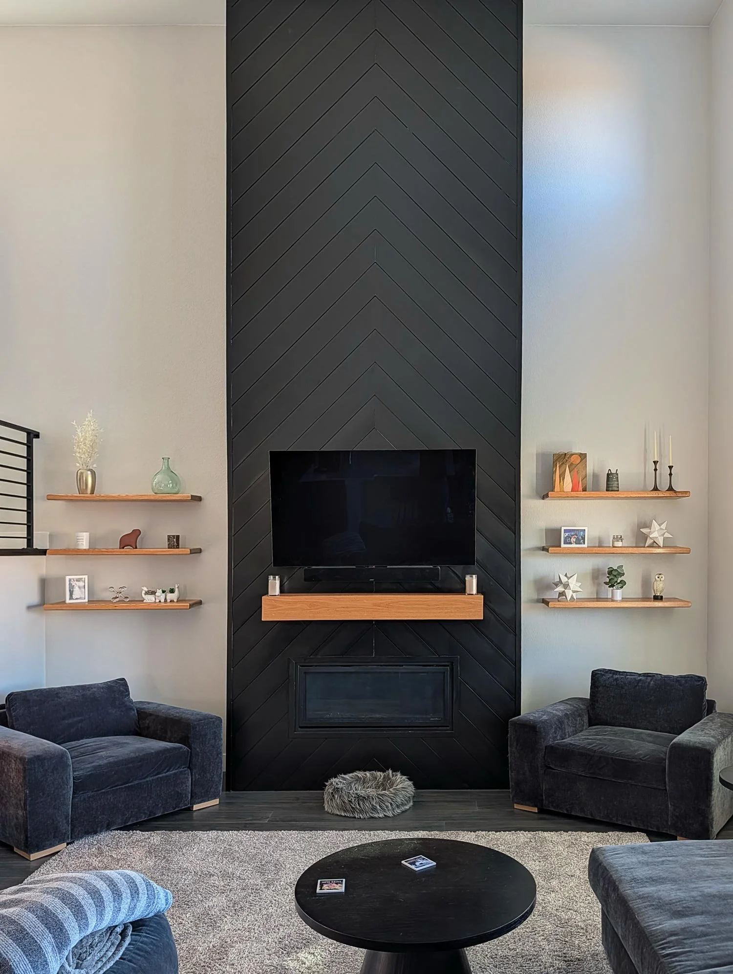 Living room with dark gray sofas, a modern black fireplace with a wooden mantel, and three floating shelves decorated with vases, books, and decorative objects. A black round coffee table with a magazine and a circular gray rug on a dark wood floor.