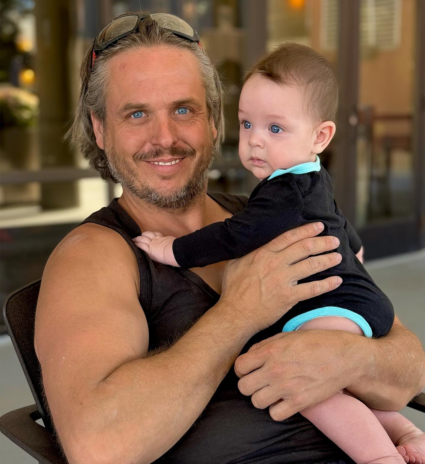 A man, holding a baby with blue eyes and light brown hair. The man is smiling and the baby has a serious expression. They are happy