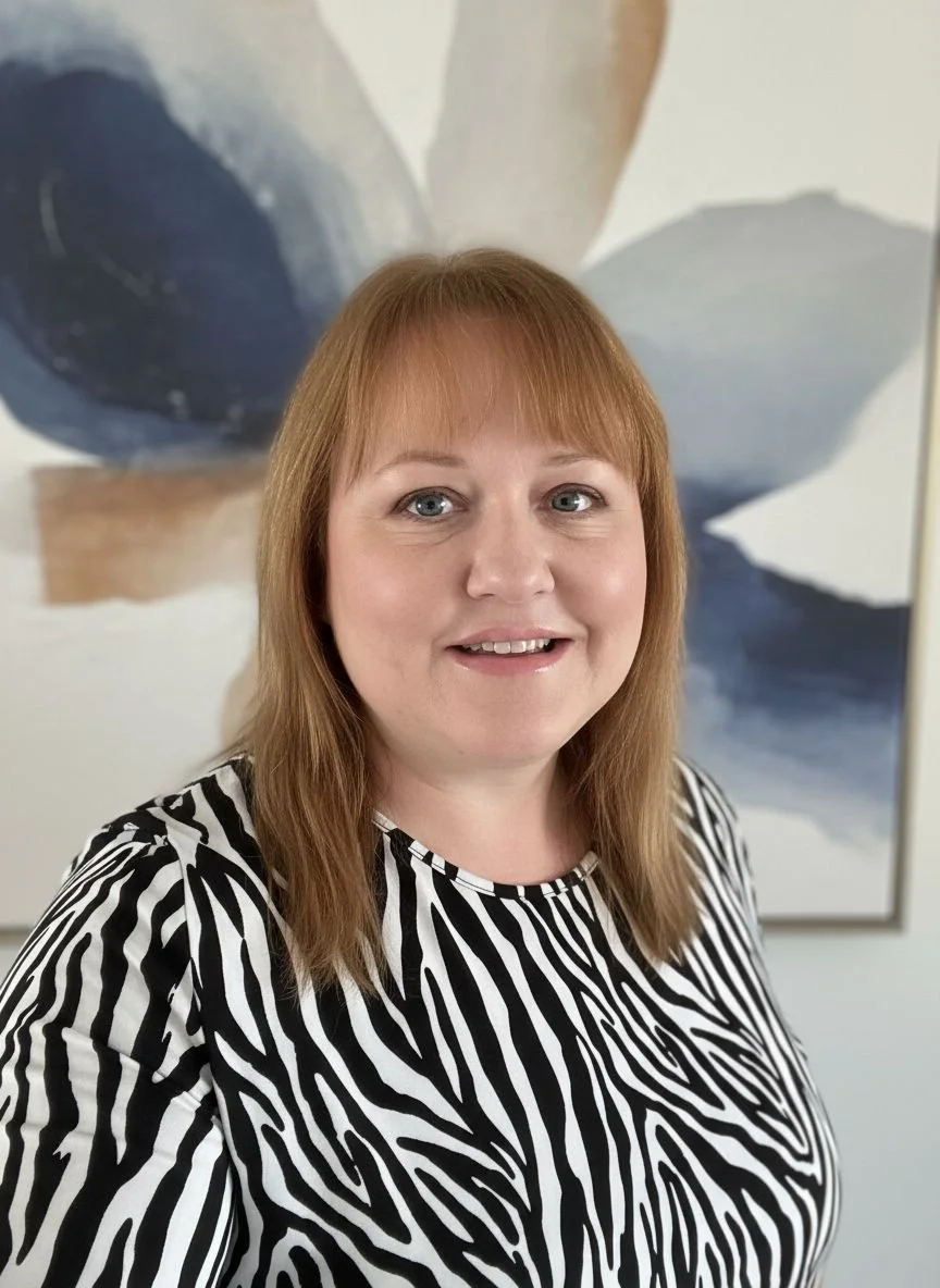 A woman with shoulder-length reddish-brown hair, wearing a black and white zebra print top, smiling in front of an abstract painting with large navy blue, beige, and white shapes.