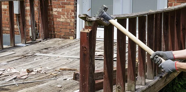 Residential deck demolition in progress with damaged wooden railing being removed using a sledgehammer during exterior renovation work.