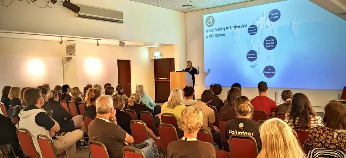Nikki Morrison presenting a slide titled 'Animal Training 101: An Overview' to an audience in a conference room with rows of attendees seated and listening.