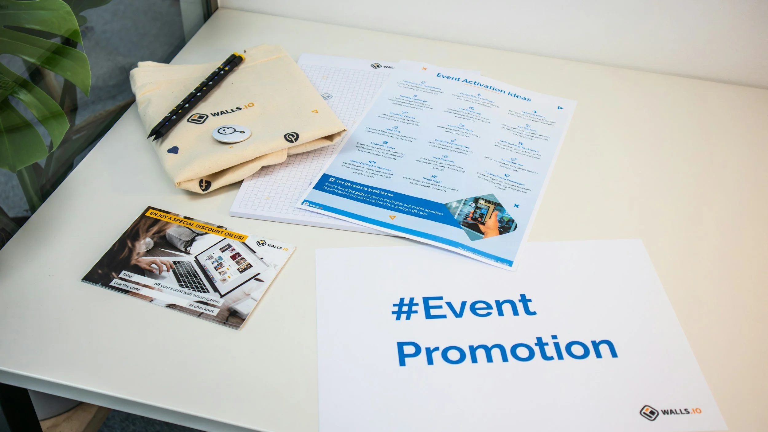 A white table with promotional materials, including a printed event promotion poster, a brochure, a fabric bag, a smiling face pin, and some pens.
