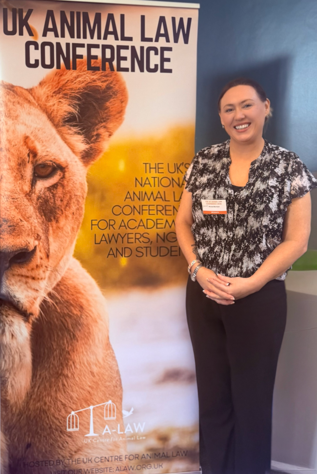 Nikki Morrison standing in front of a banner for the UK Animal Law Conference. The banner features a close-up image of a lion cub and the event details.