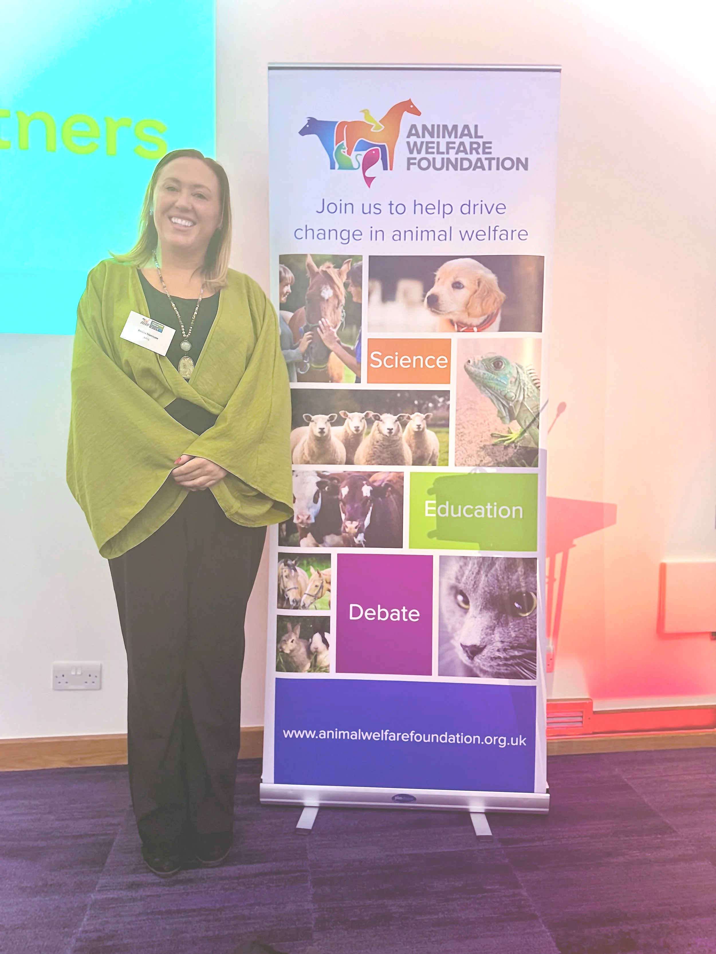 Nikki Morrison standing next to an Animal Welfare Foundation banner with images of various animals including a dog, a cat, sheep, a horse, and wildlife, with the foundation's website URL at the bottom.