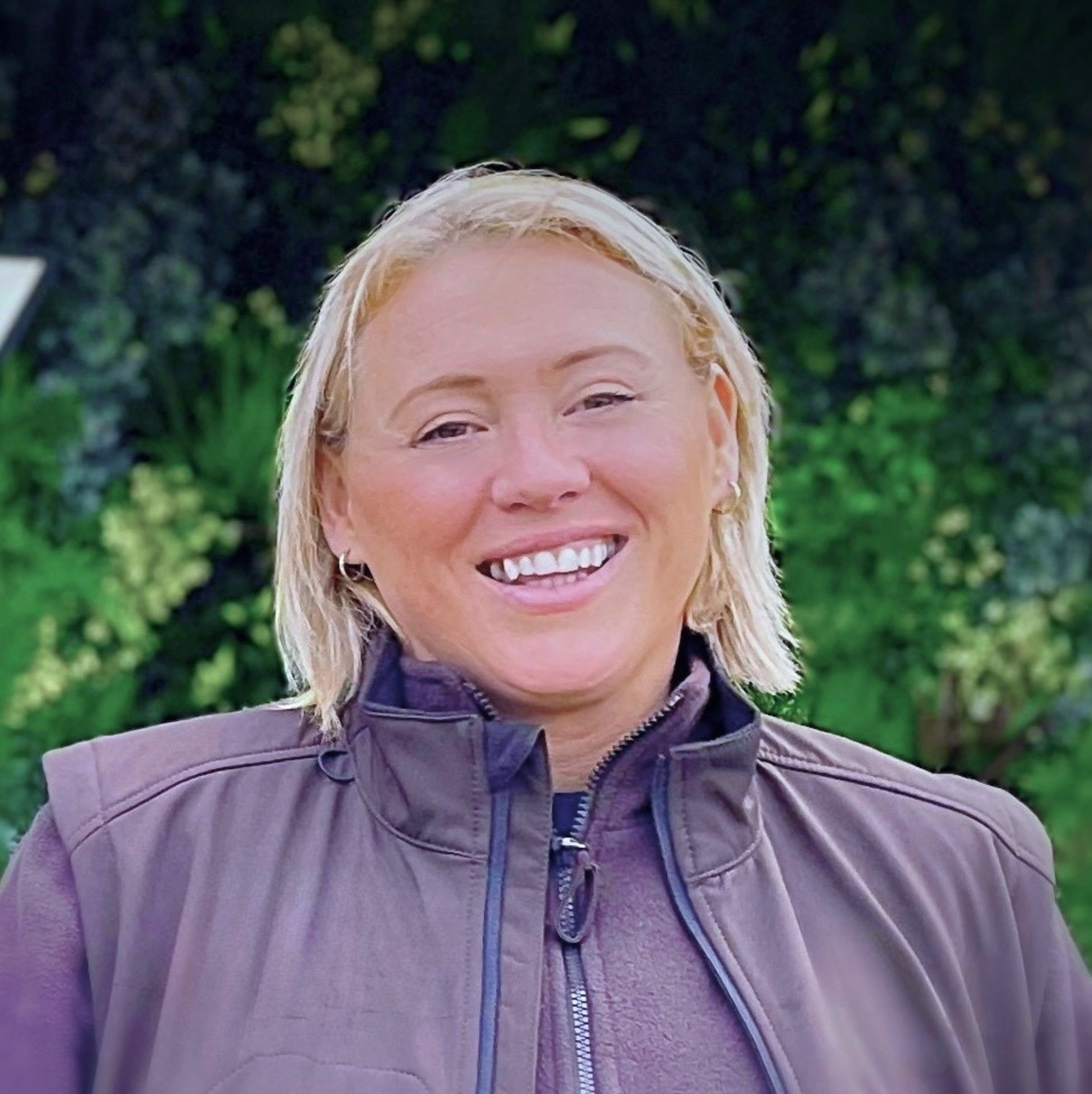 Close-up of Nikki Morrison smiling, wearing a dark jacket with a zipper, standing outdoors with greenery in the background.