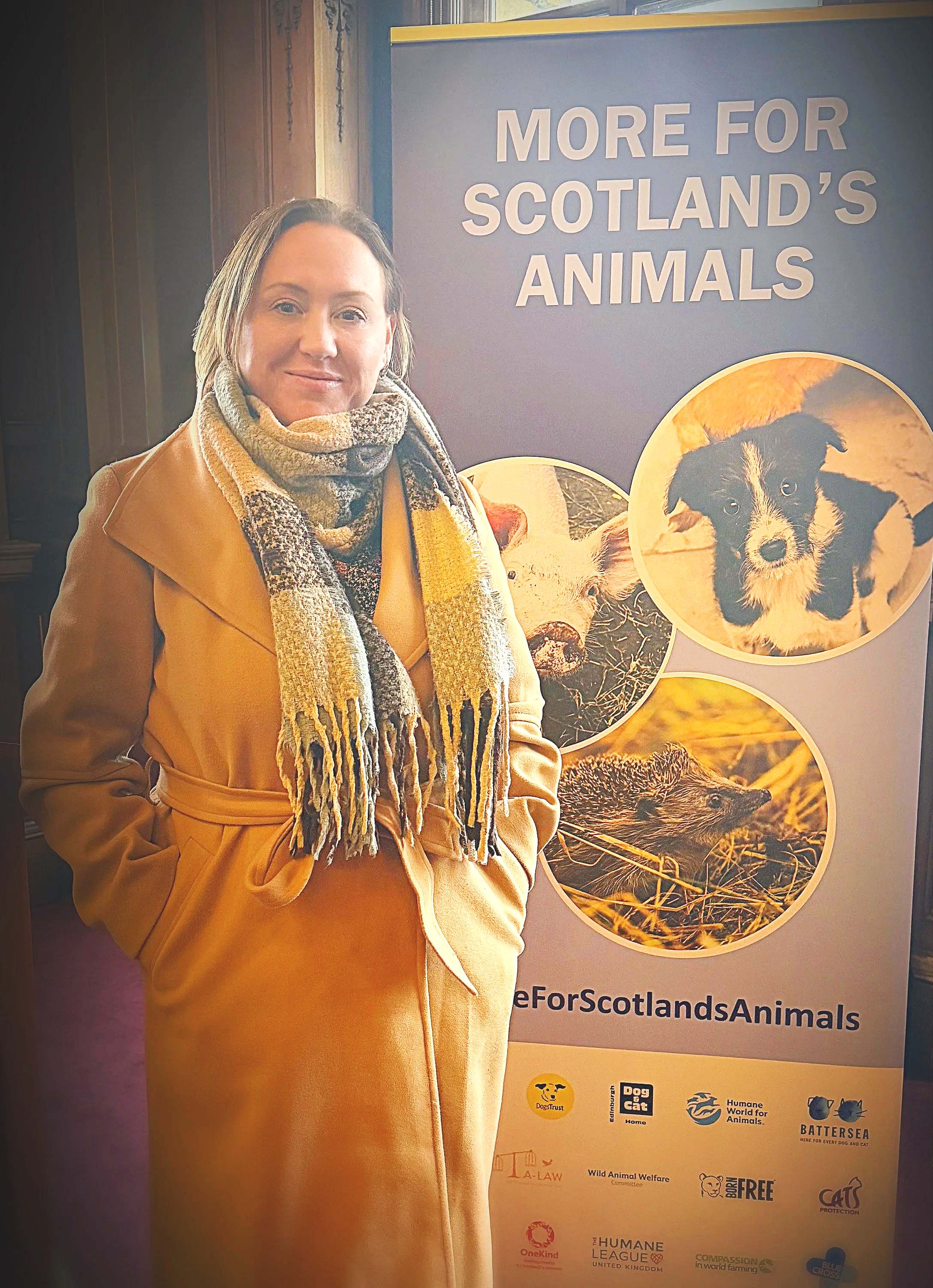 Nikki Morrison standing next to a banner that reads 'More for Scotland's Animals' with images of various animals, and logos of organisations such as Dogs Trust, Humane World for Animals, Battersea, and Born Free.