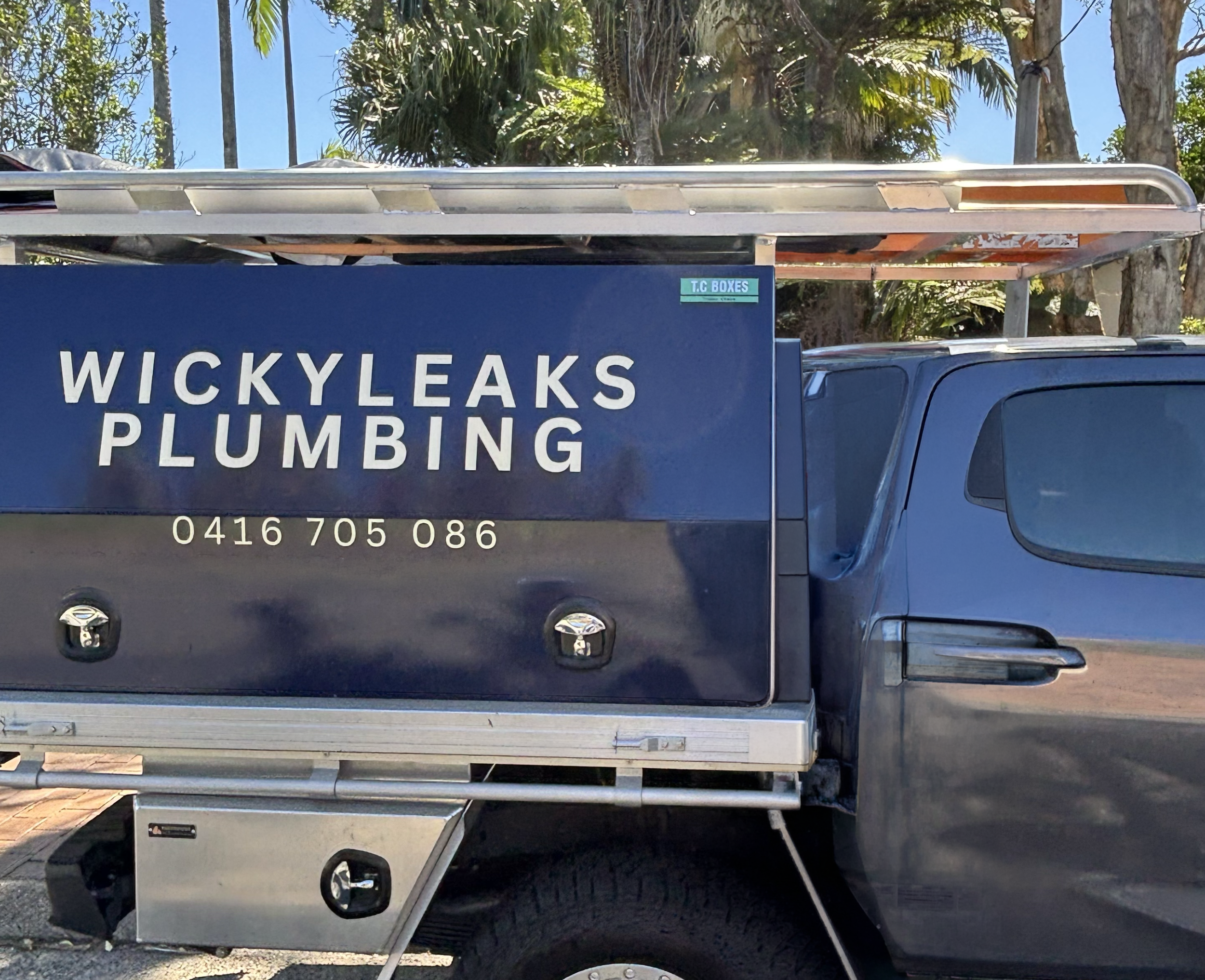 A blue van with white lettering advertising WICKYLEAKS PLUMBING and phone number 0416 705 086. The van has a sign on the side that reads 'T.C BOXES.' The background includes trees and a clear blue sky.
