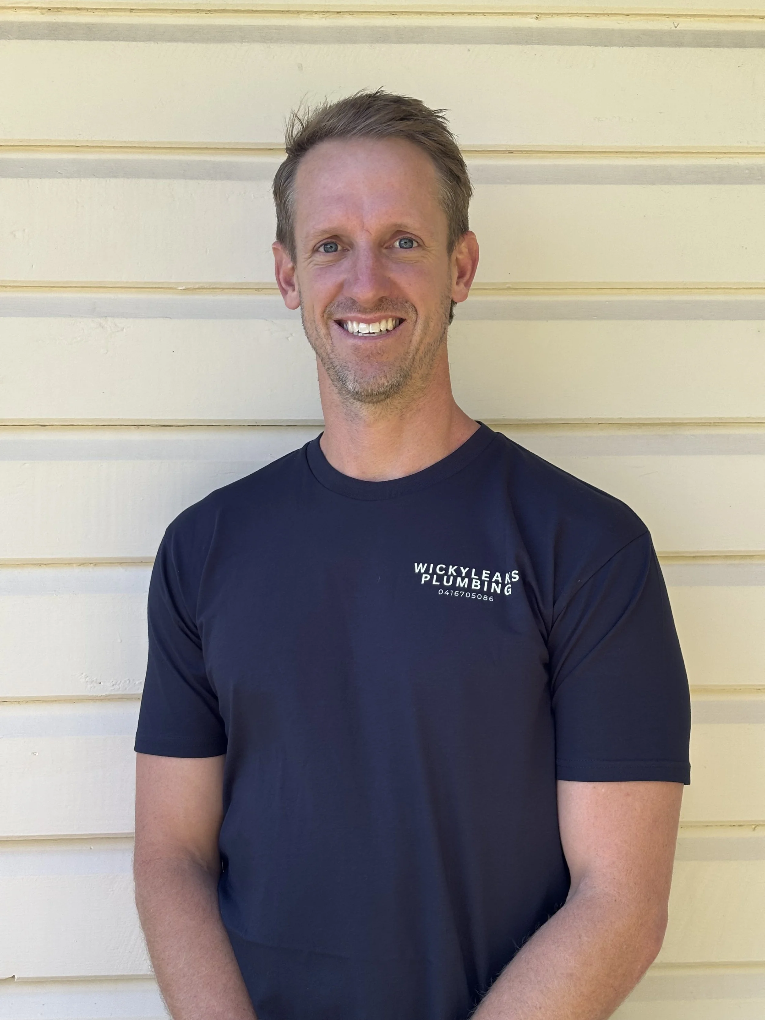 A smiling man with blue eyes and short brown hair wearing a black t-shirt with a plumbing company logo, standing in front of a cream-colored wooden wall.