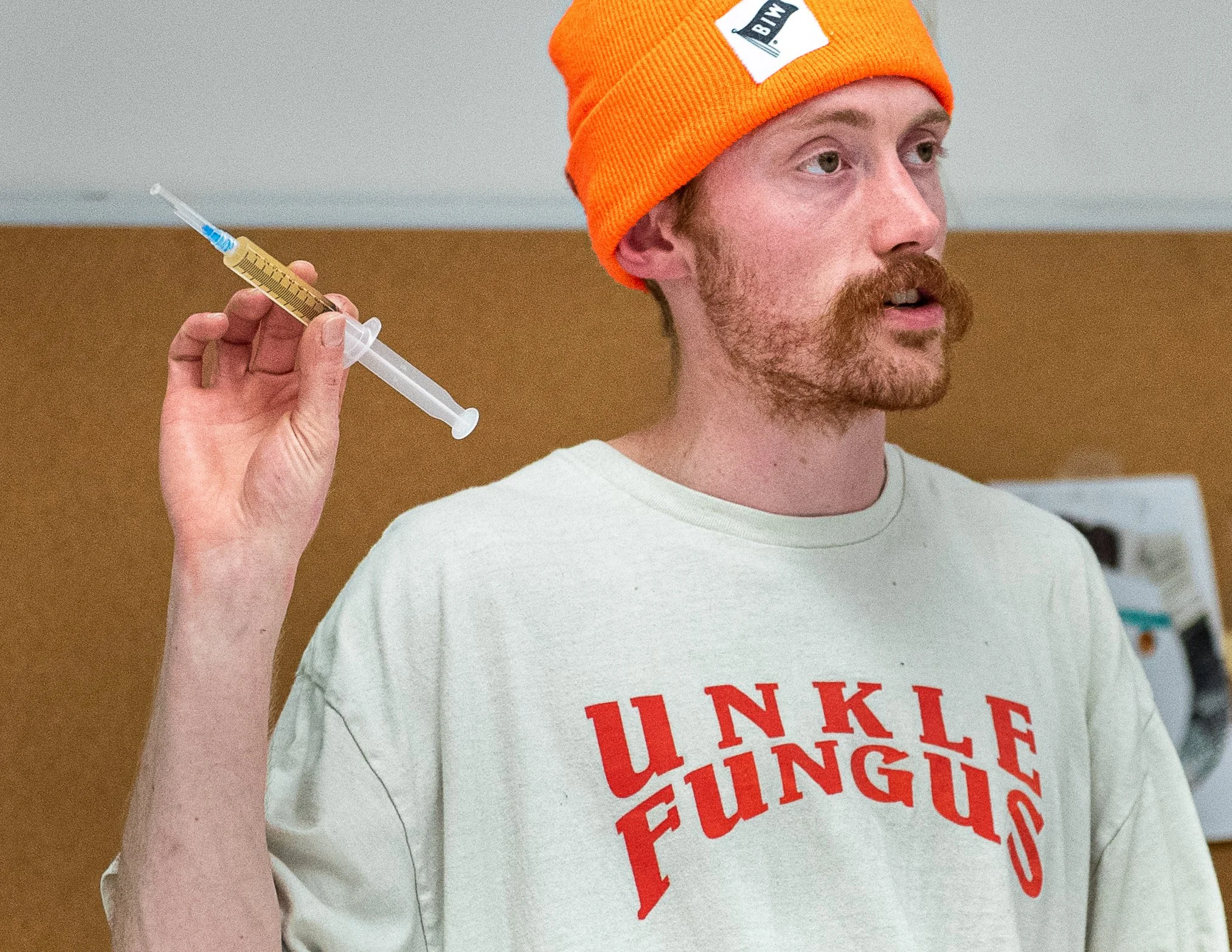 A man wearing an orange beanie hat with a BMW logo, holding a medical syringe near his face, wearing a white T-shirt with red text.