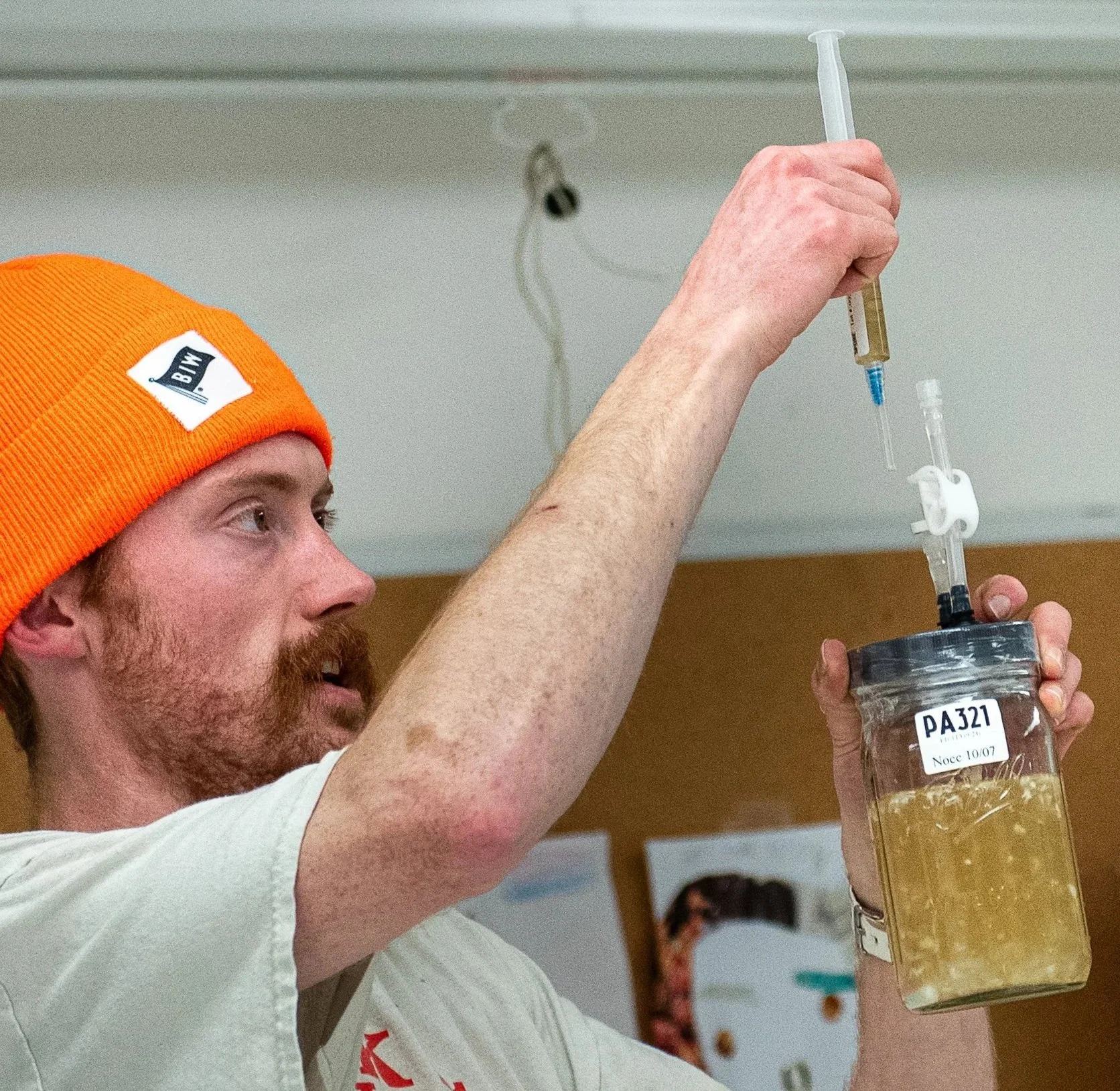 A man with a beard and orange beanie preparing a syringe, with a vial of medical liquid in hand, likely for vaccination or injection in a clinical setting.