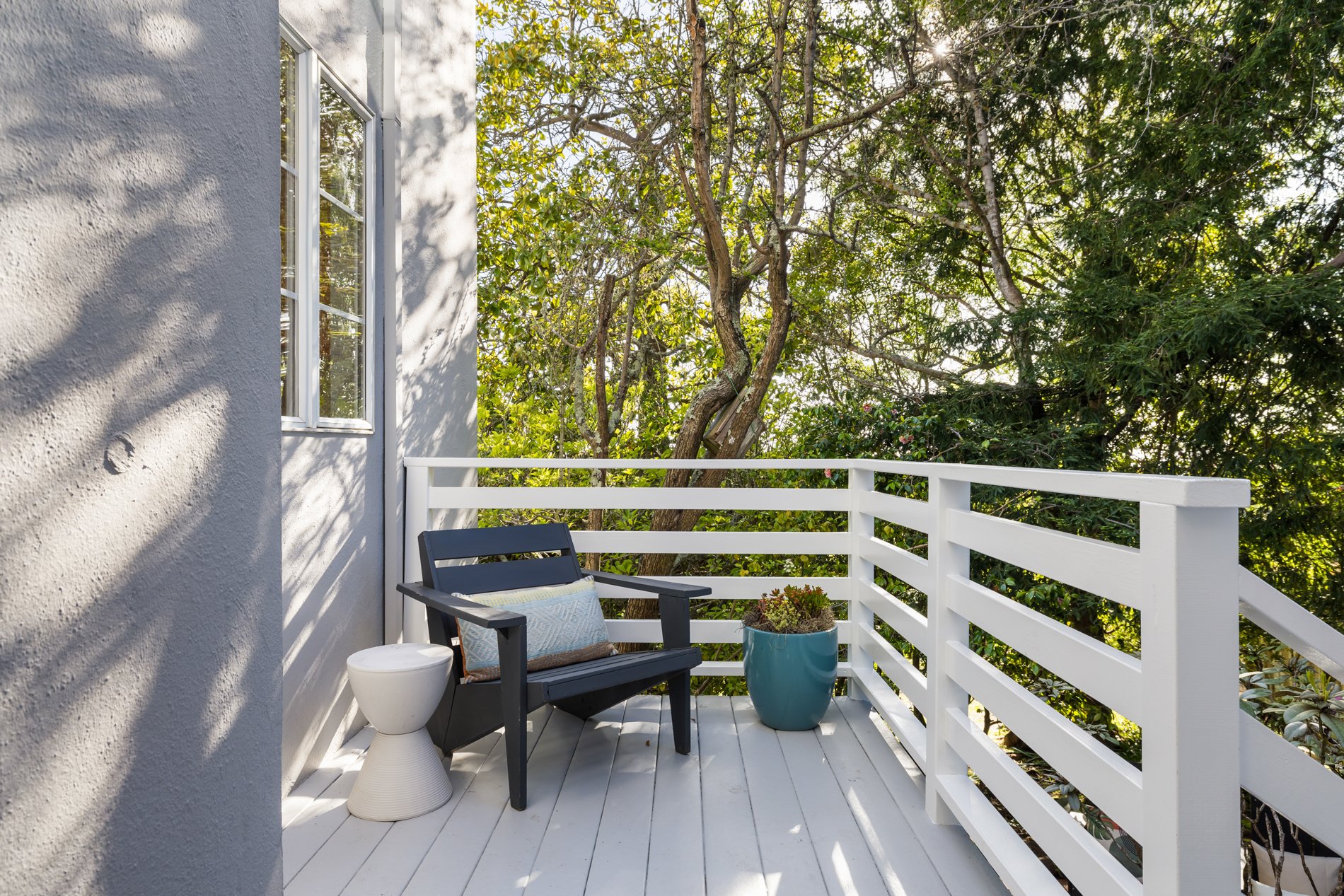 Small white balcony with white railing, black chair with cushion, potted plant, toilet, and window, surrounded by trees and greenery.