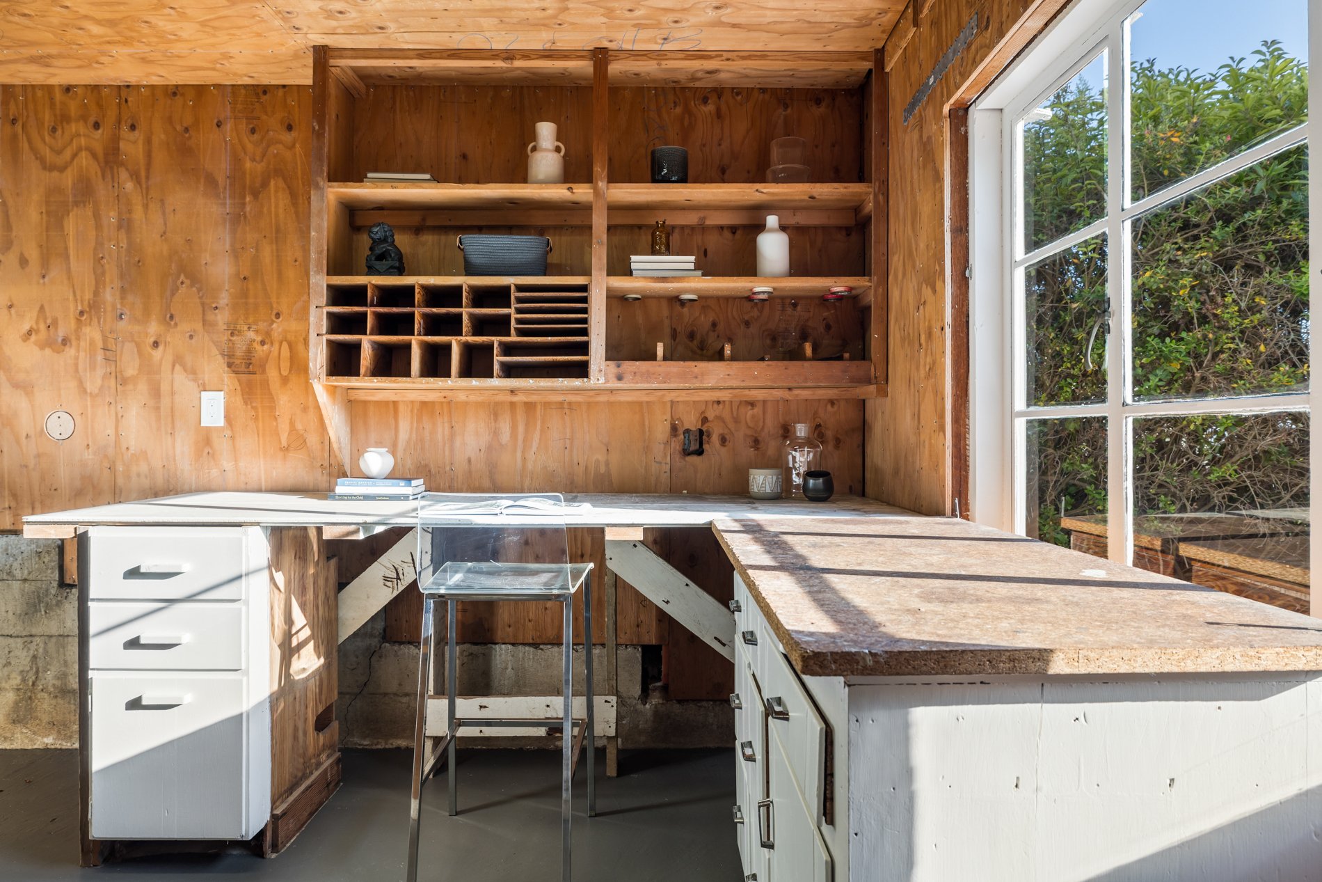 Interior view of a rustic kitchen with wooden wall shelving, a window showing greenery outside, a white cabinet with drawers, a small clear chair, and various decorative items on the shelves.