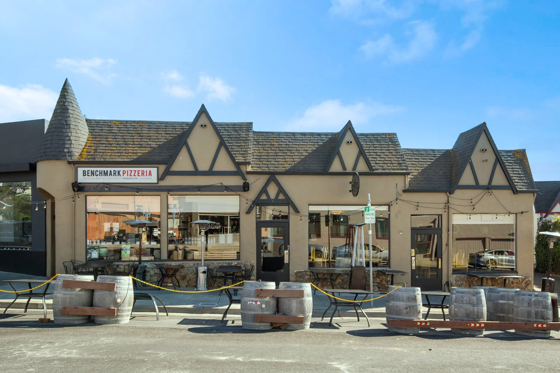 Exterior view of a restaurant named Benchmark Pizzeria with Tudor-style architecture, hanging string lights, outdoor seating, and a parking lot in front.