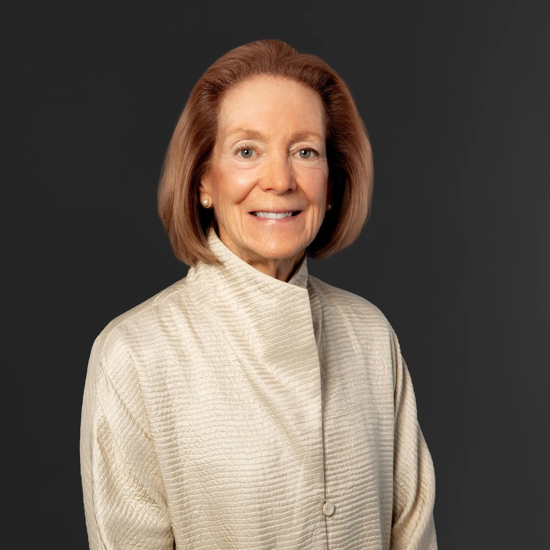 A portrait of an older woman with short, light brown hair, wearing a beige textured shirt with a high collar, smiling against a dark gray background.