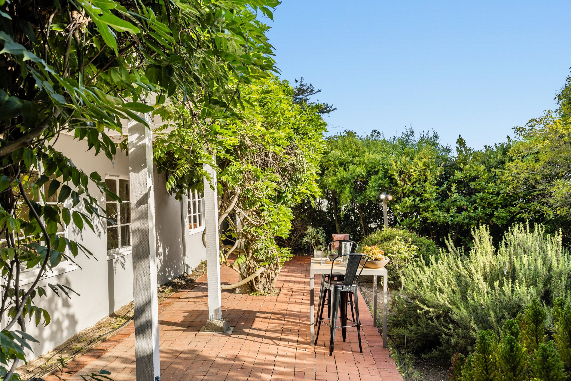 A sunny backyard with a brick patio, outdoor seating and lush green trees and plants surrounding the area.