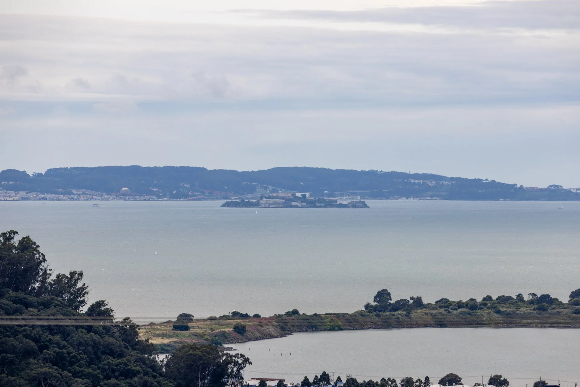 A view of a harbor with water, islands, and hills in the background, under a cloudy sky, with trees in the foreground.