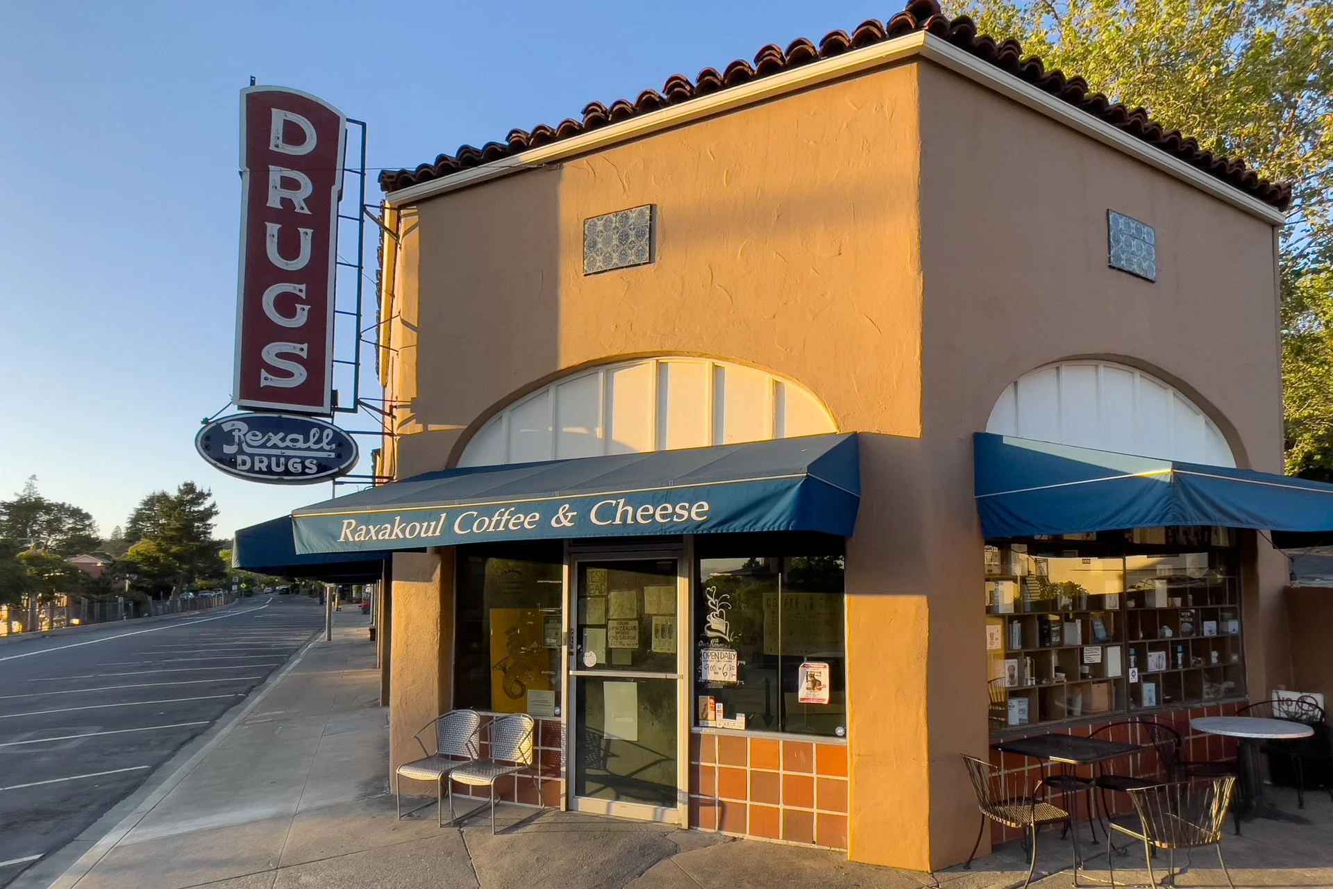 Outside of a building with a sign that says "DRUGS" and a smaller sign below that says "Rexall Drugs." There is a storefront with a blue awning that reads "Raxakoul Coffee & Cheese". There are chairs and tables outside the store. The building has a b