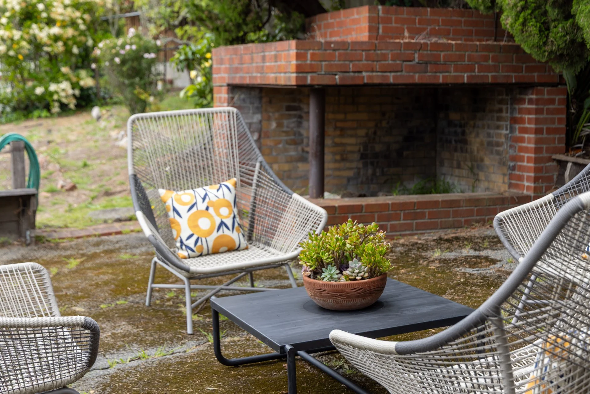 Outdoor patio with metal chairs, a table with a potted succulent, and a brick fireplace in a garden setting.