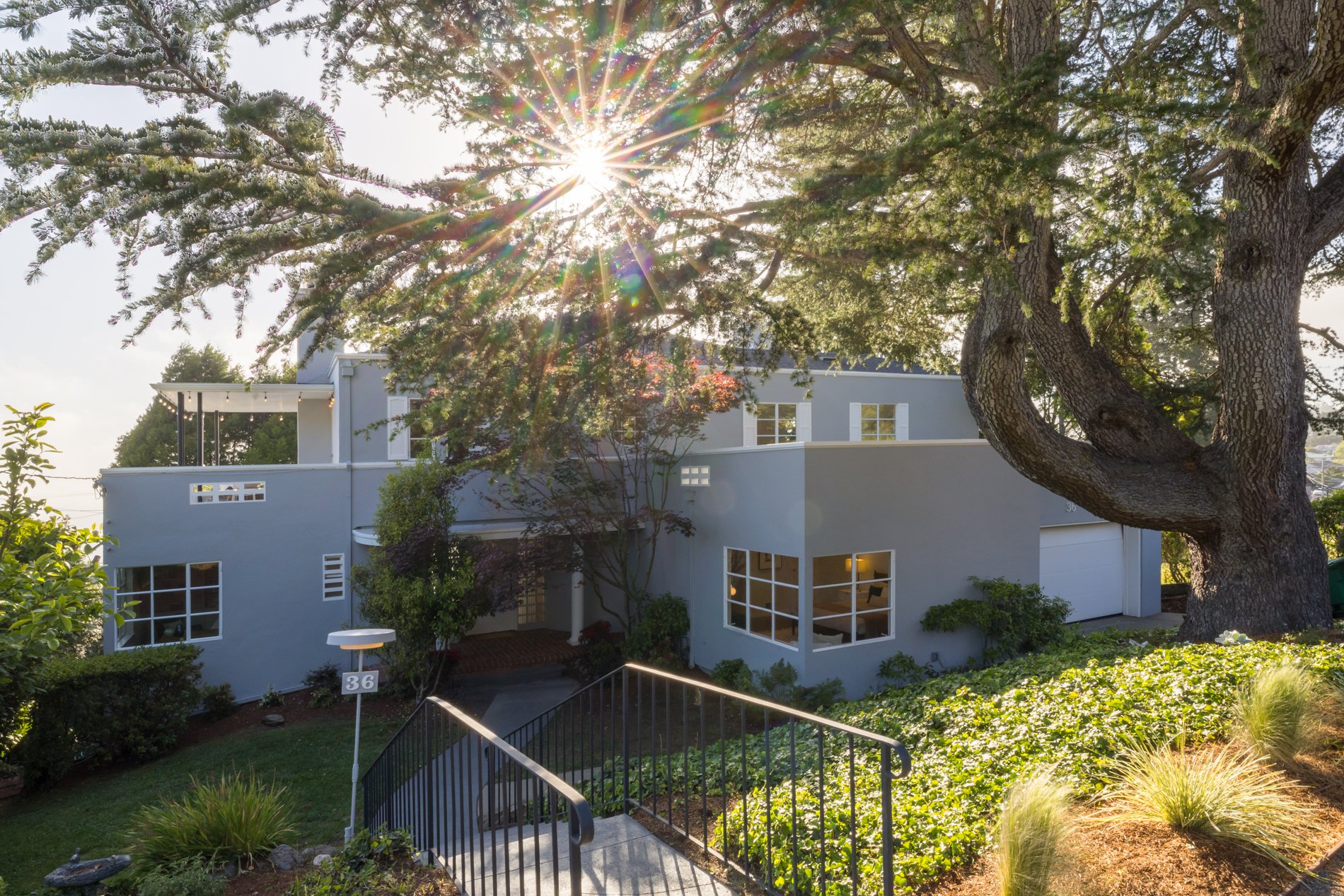 A modern, multi-story house painted light gray with white window frames, front porch lighting, and a garage, surrounded by lush greenery, including a large tree with sprawling branches partially blocking sunlight.