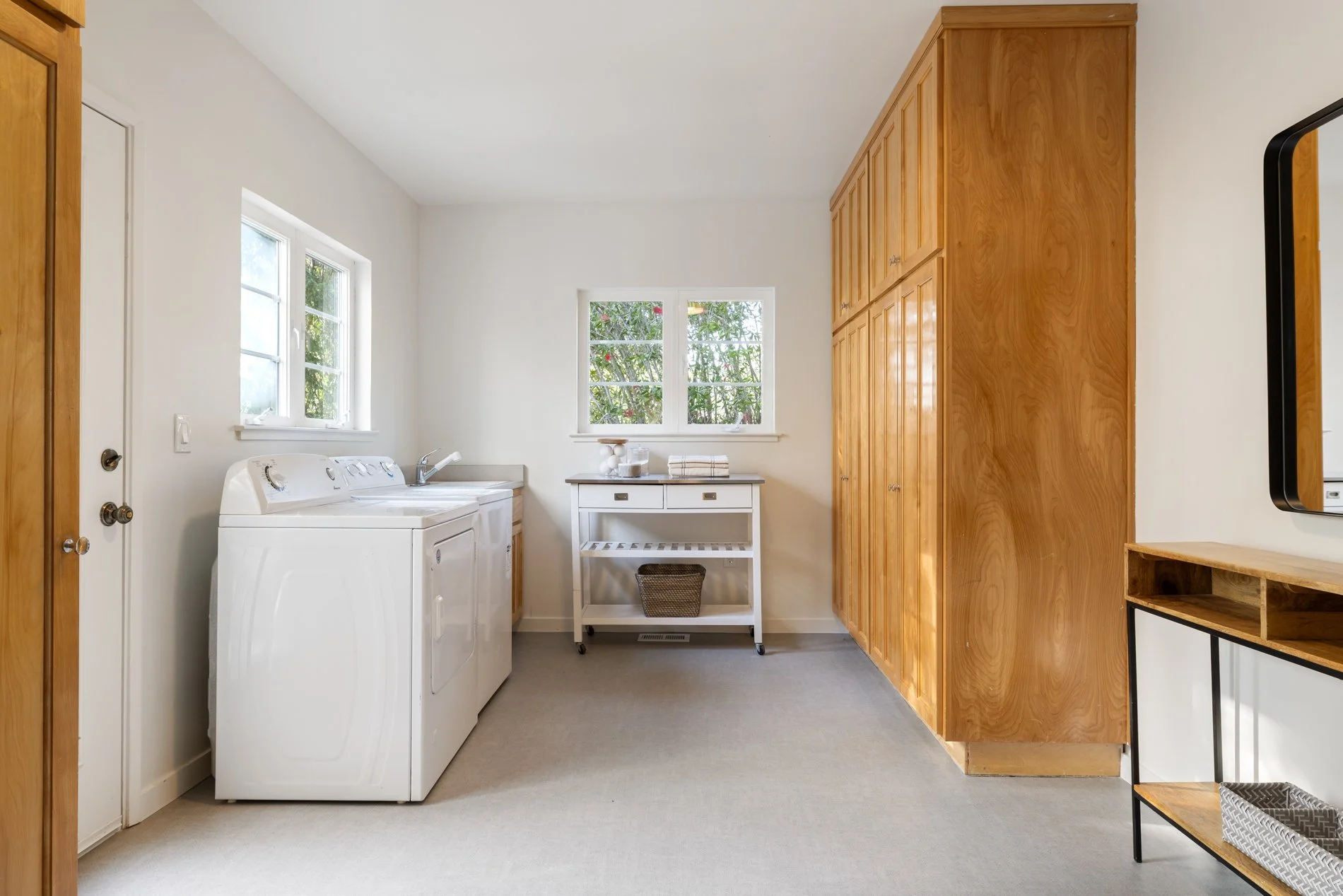 A laundry room with white walls, two windows, a white washing machine and dryer, a small white table with folded towels, wood cabinet storage, and a mirror on the wall.