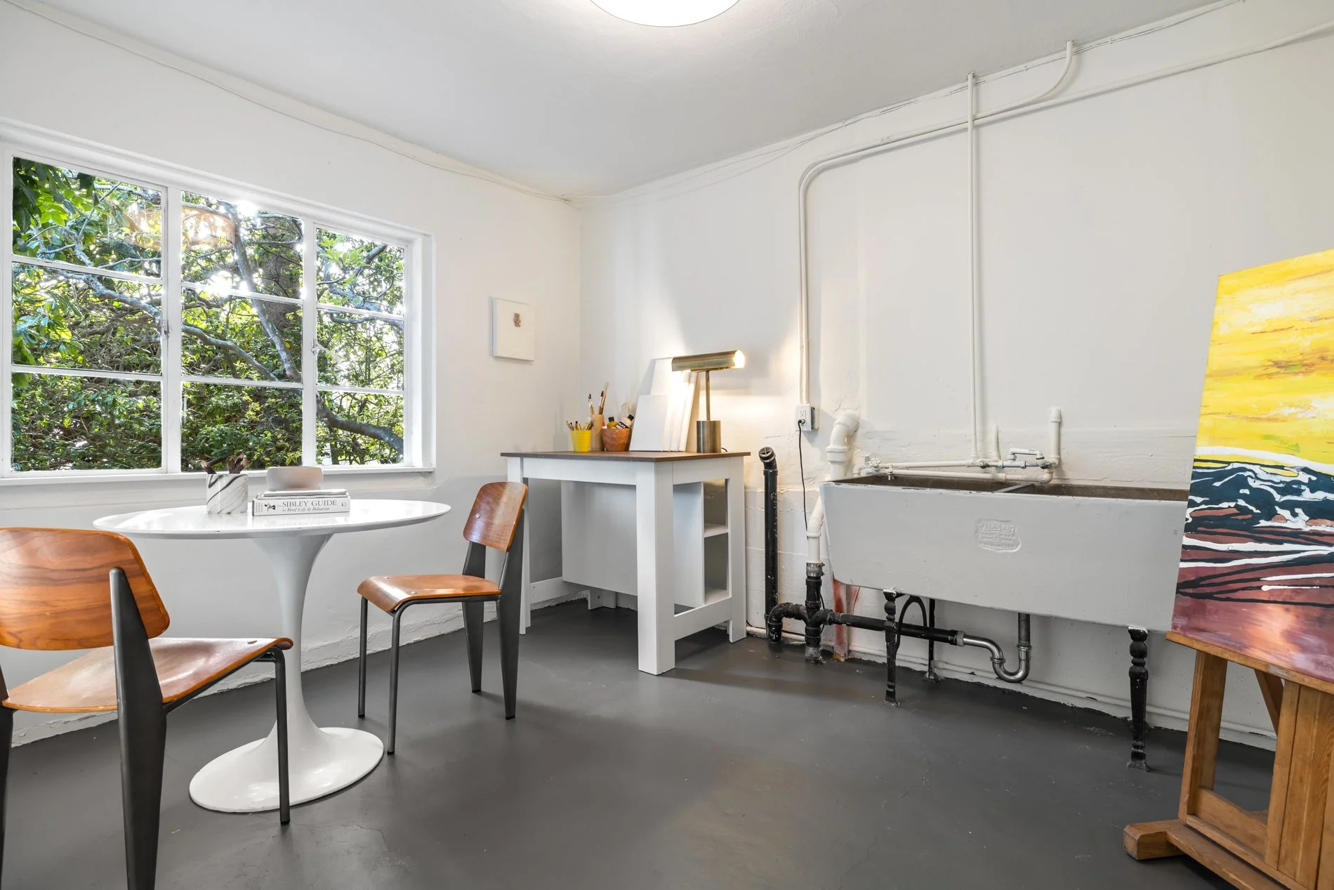 Interior of an art studio or cafe with a large window, round white table, wooden chairs, artwork, and art supplies. There is a utility sink against the wall.